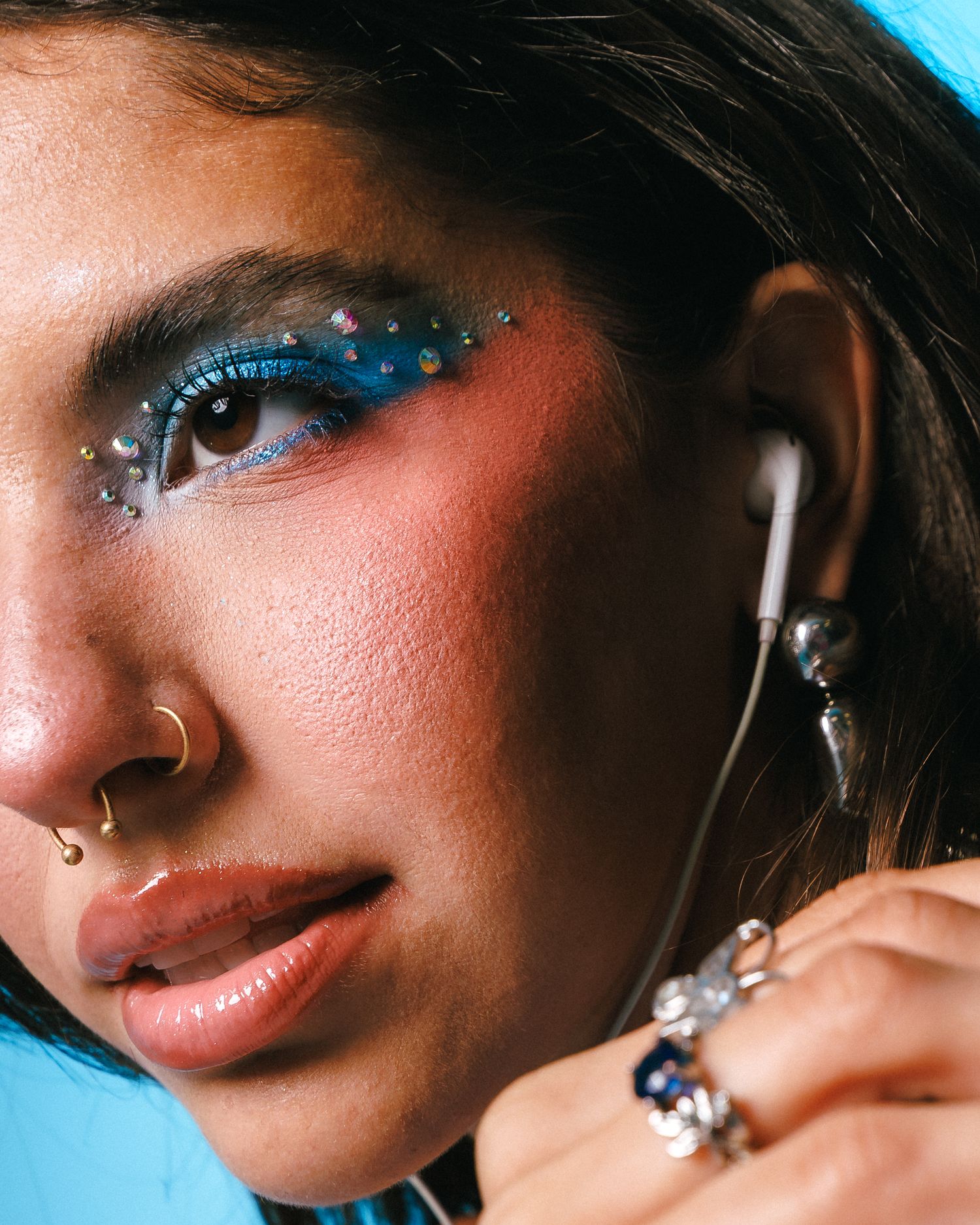 Close-up of a woman with blue glitter eye makeup, rhinestones, rosy blush, nose ring, rings, and earbuds against a blue background.