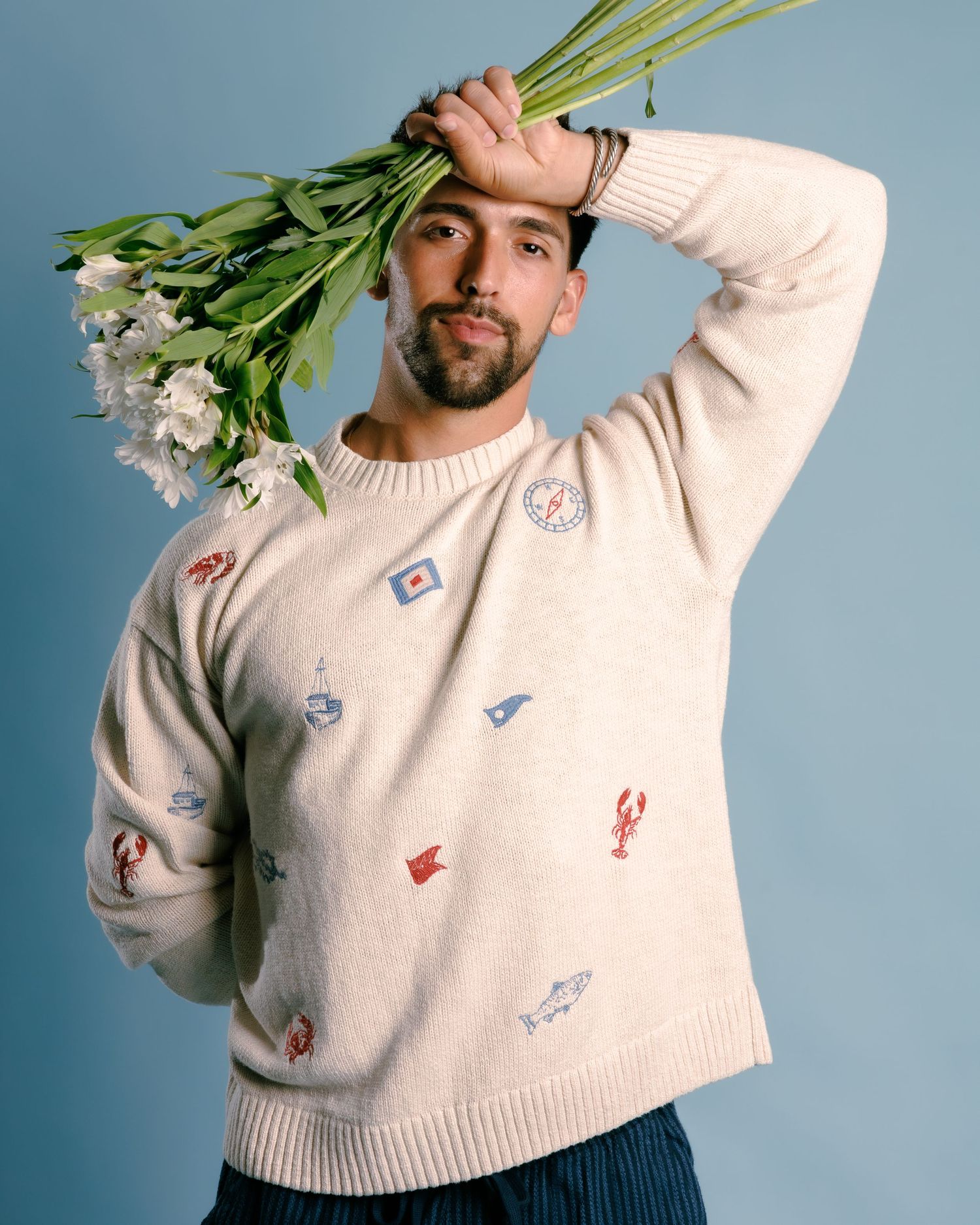 Man in cream nautical embroidered sweater holding white flowers above his head against a blue background.