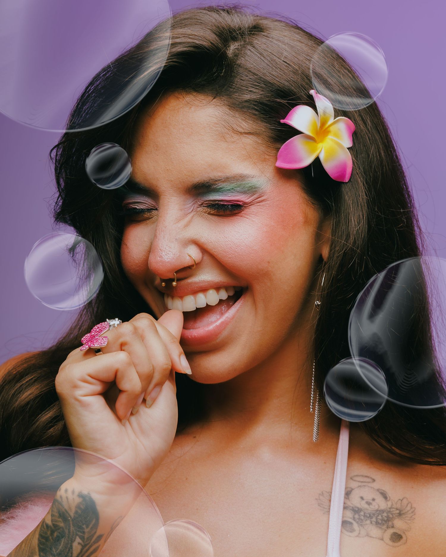 Smiling woman with colorful eye makeup, pink flower in hair, nose ring, and tattoos against a purple background with bubbles.