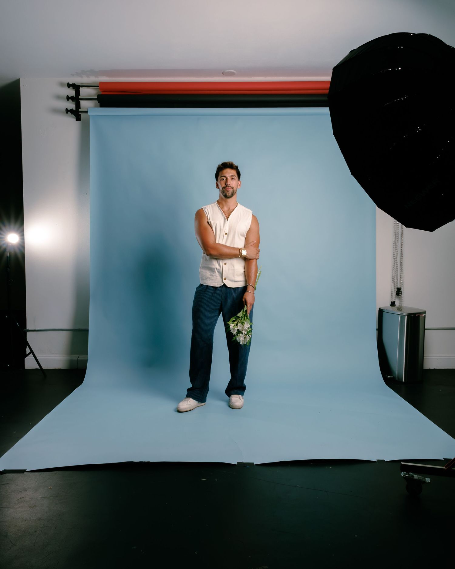 Man in white sleeveless shirt and navy pants holds flowers, posing in photography studio with blue backdrop.