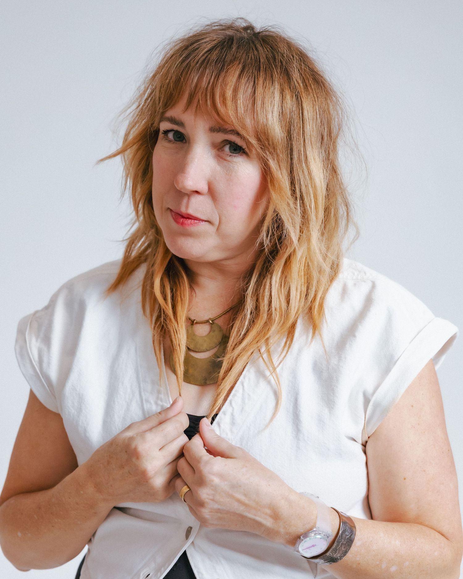 Portrait of blonde woman with bangs wearing white top adjusting collar against gray background.