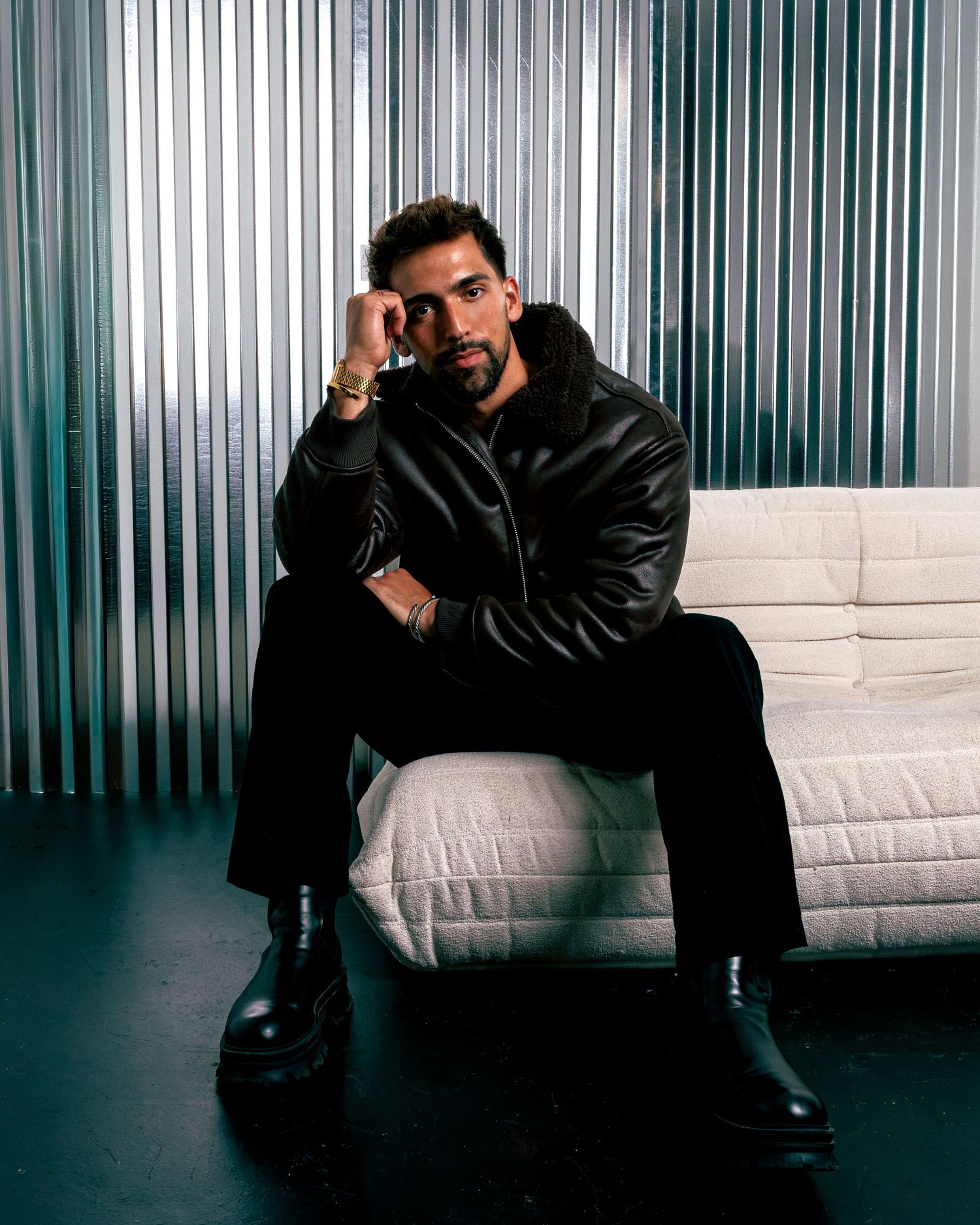 A stylish man in a black leather jacket and trousers sits on a white sofa against a metallic backdrop.