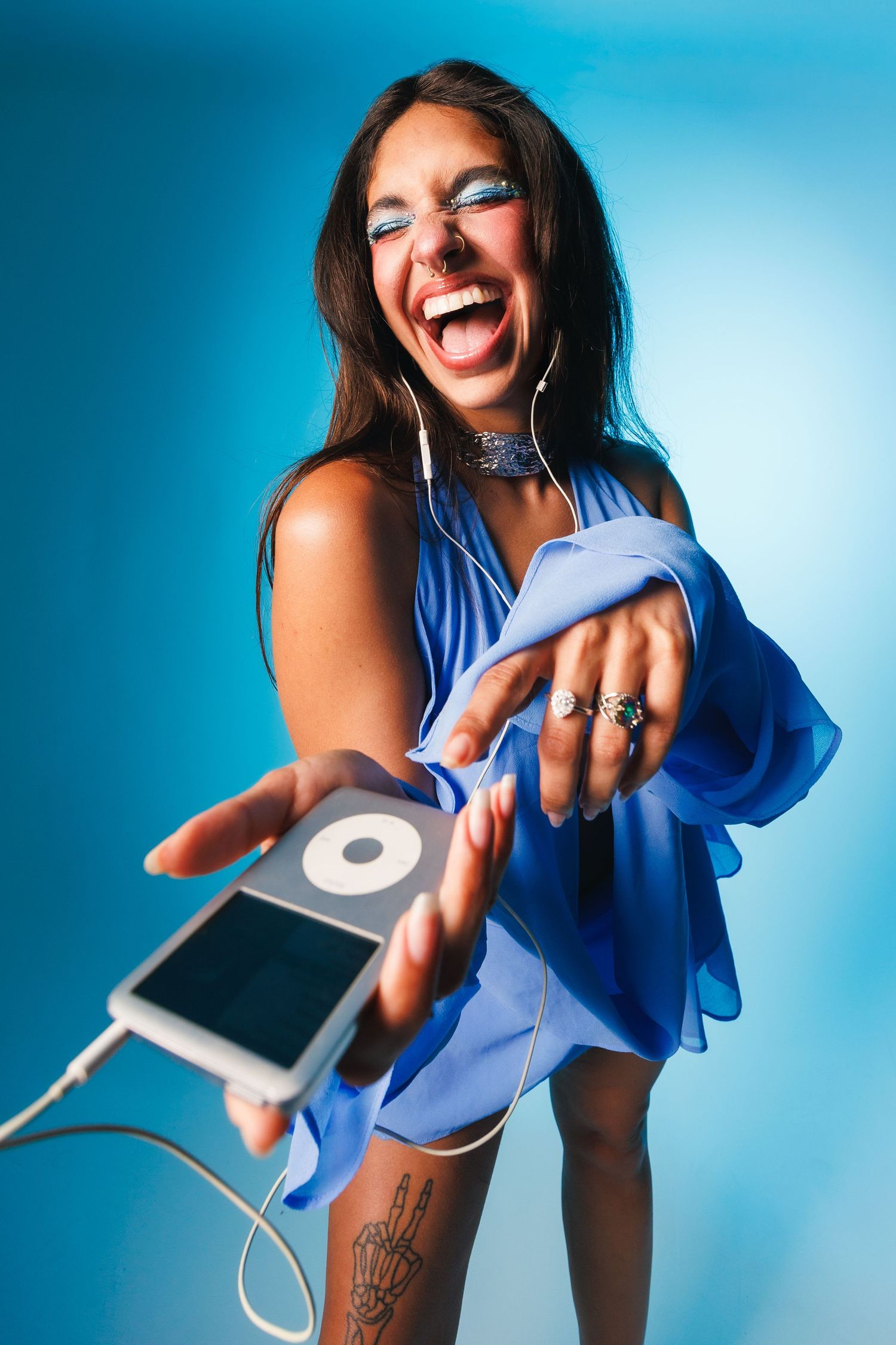 Laughing woman in blue dress holding iPod classic with earbuds, blue makeup, skeleton hand tattoo, blue studio background.