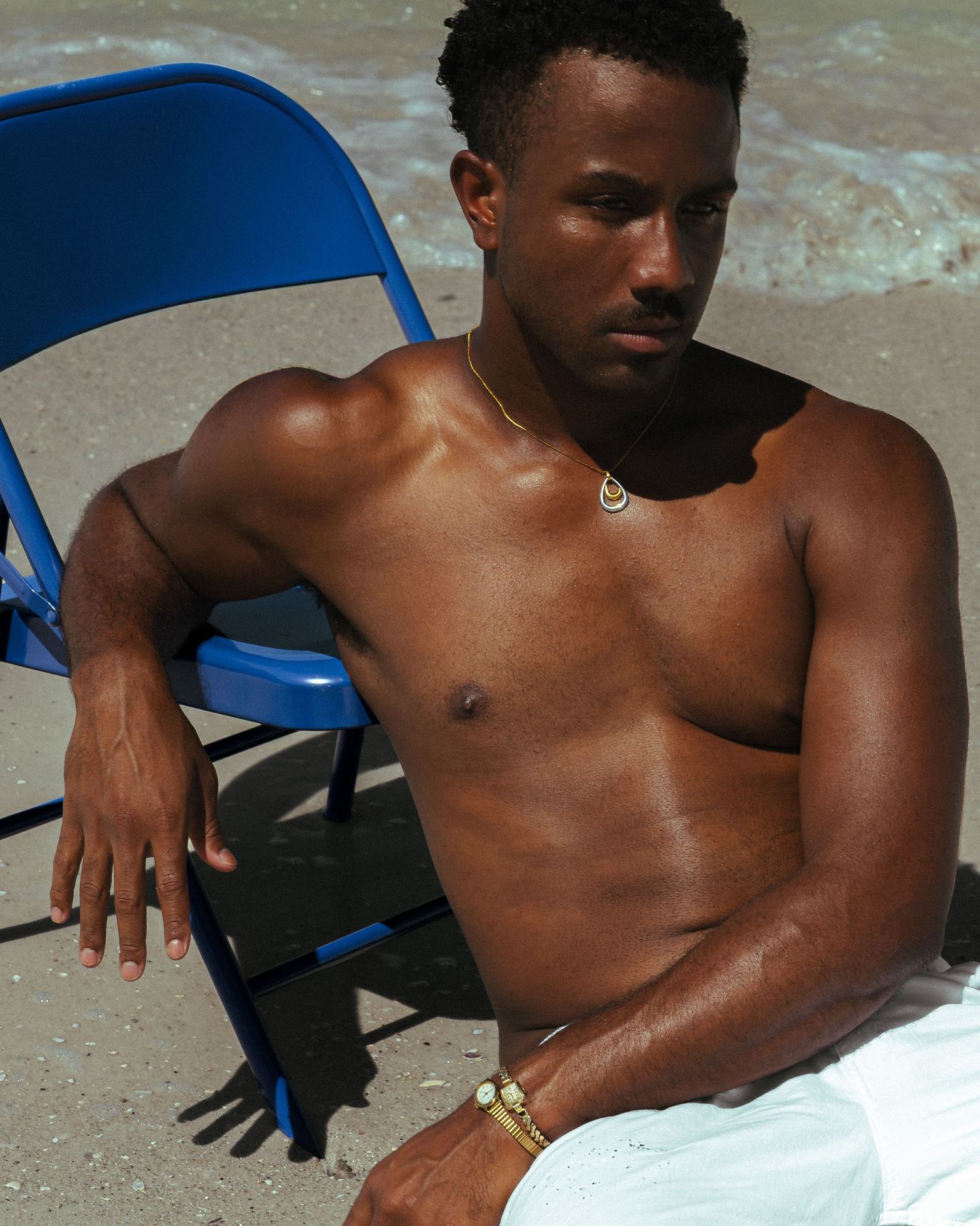 Shirtless man wearing gold jewelry relaxes on a blue folding chair at the beach on a sunny day.