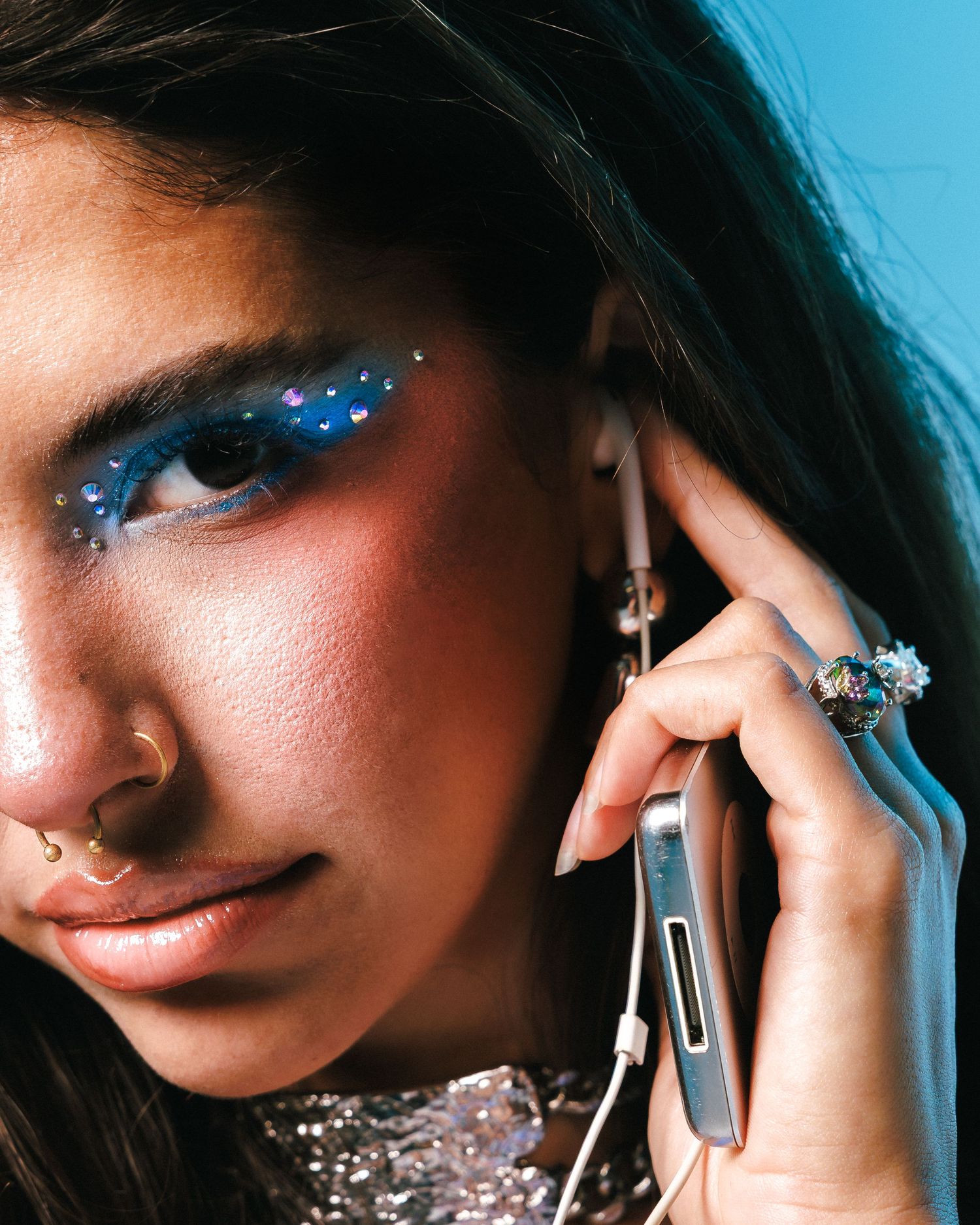 Woman with glittery blue eye makeup holds phone to ear, wearing nose ring and jeweled ring against blue background.