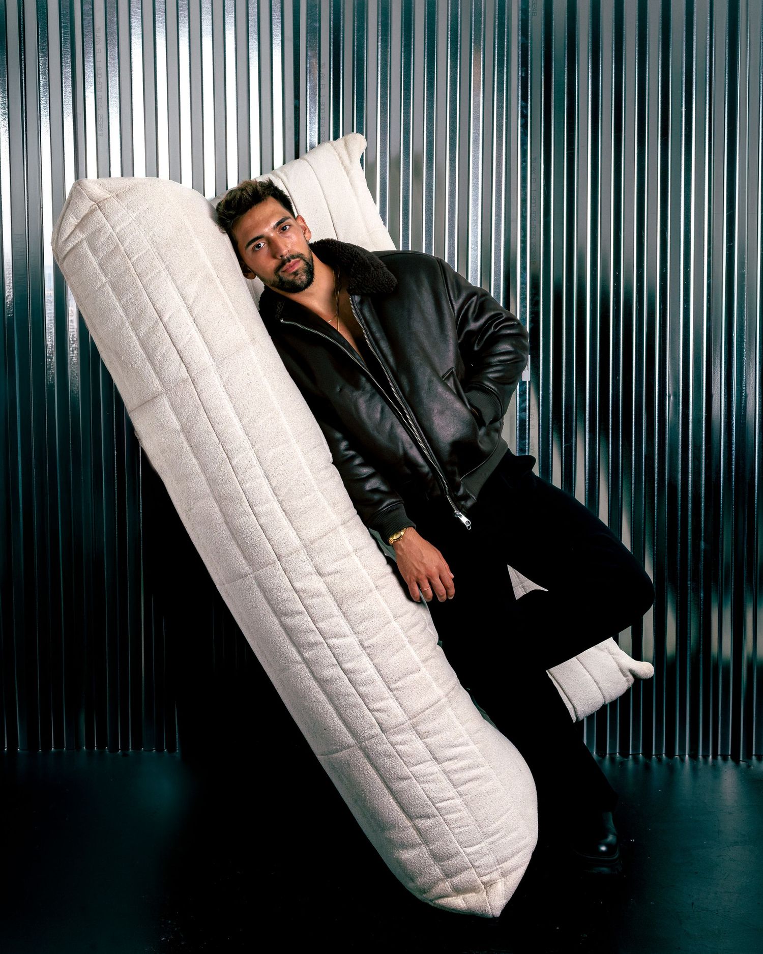 Man in black leather jacket posing with oversized quilted body pillow against metallic corrugated background.