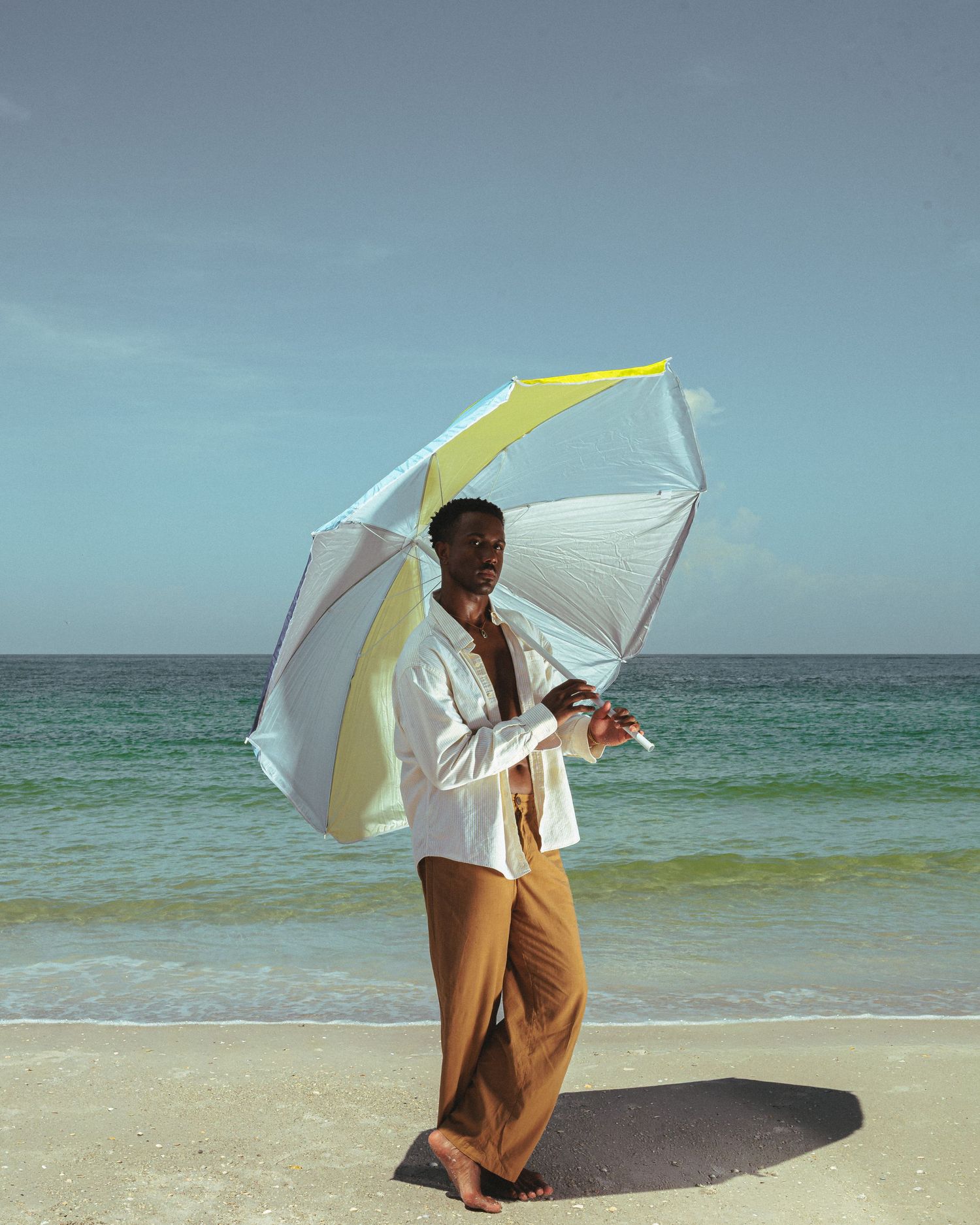 A man stands on a tropical beach holding a yellow and white umbrella, wearing a white shirt and tan trousers.
