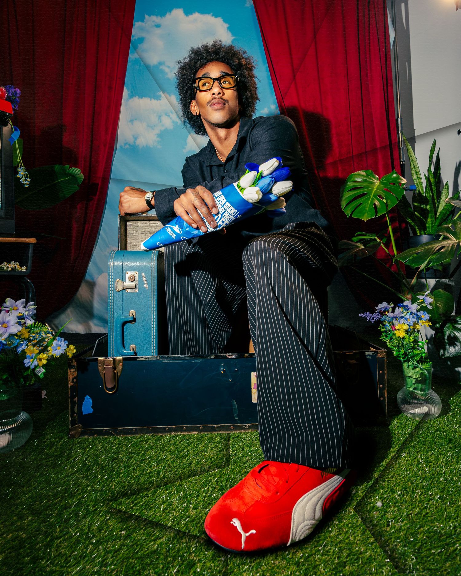 Man in pinstripe suit sitting on vintage luggage wearing oversized red Puma slippers, holding blue sock, surrounded by flowers.