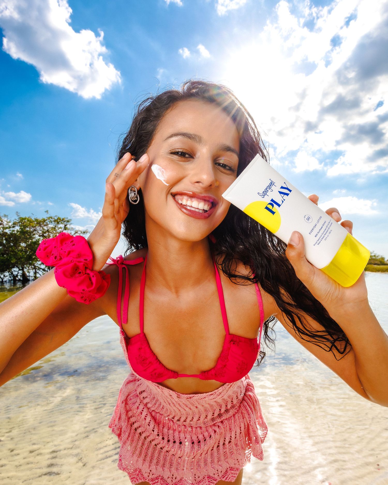 Smiling woman in pink bikini applying Supergoop PLAY sunscreen at sunny beach.