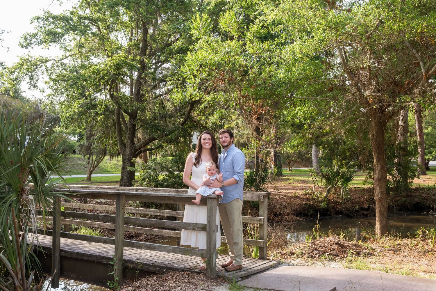 A couple stands on a wooden bridge holding their baby, surrounded by lush green trees and a serene pond.