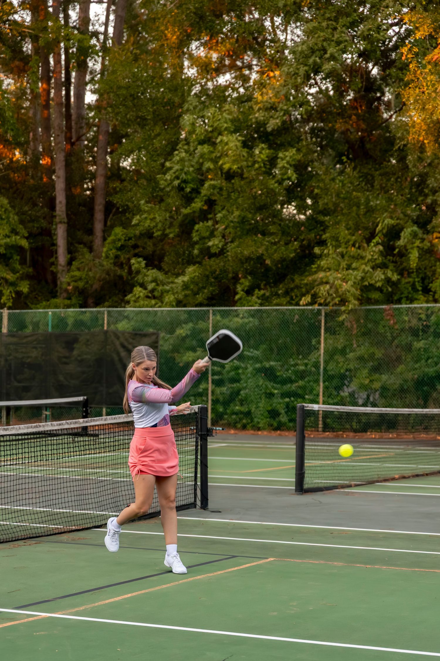 Person playing pickleball on outdoor court surrounded by autumn trees.