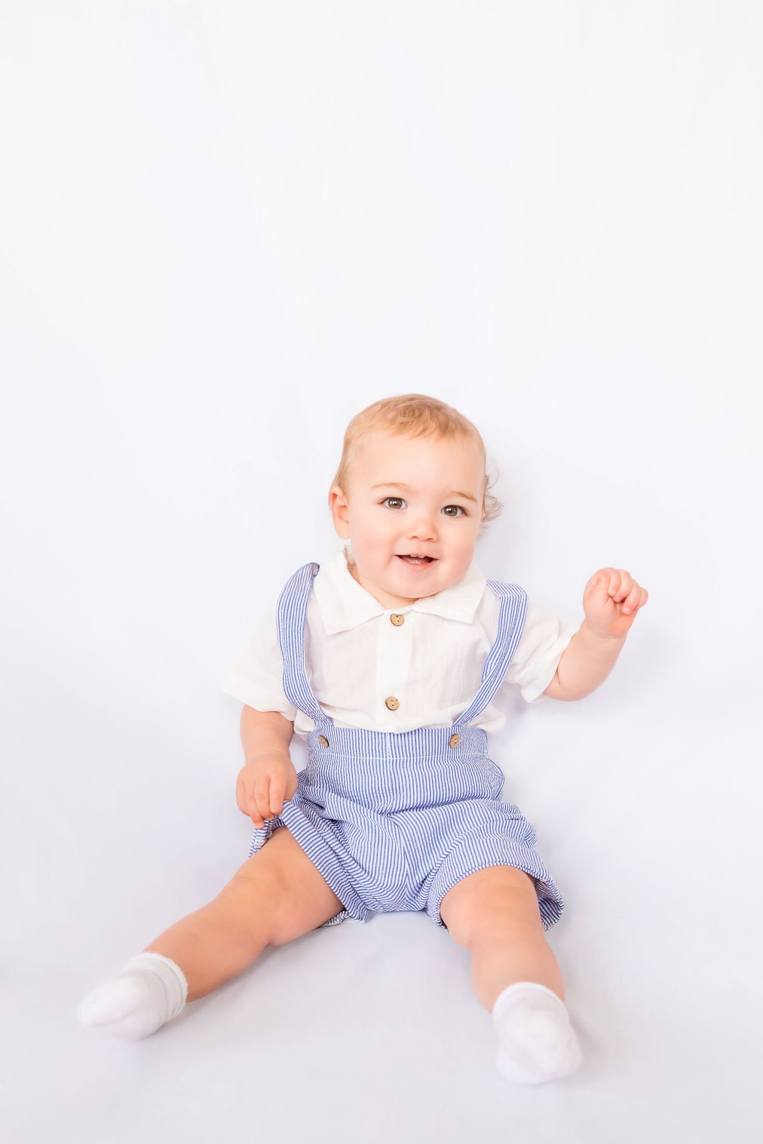 Adorable baby boy in blue overalls and white shirt sitting on white background, smiling happily.