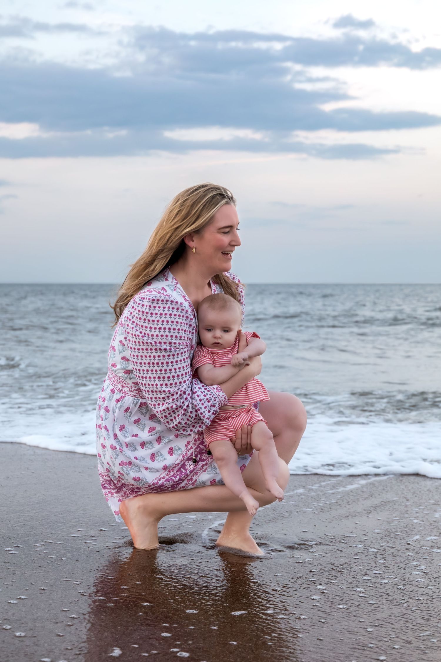 A smiling mother crouches at ocean's edge holding her baby in pink outfit as gentle waves wash ashore at sunset.