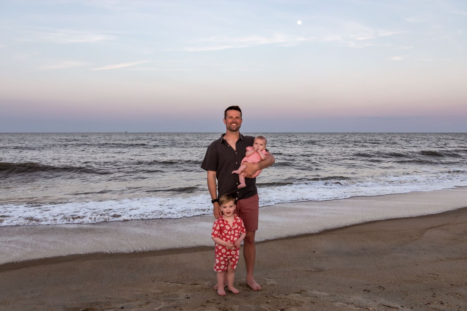 Man holding baby with toddler girl standing beside him on a sandy beach at sunset with calm ocean waves.