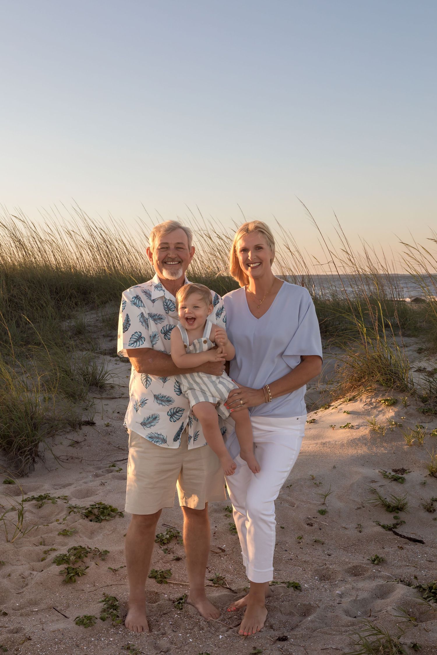 Grandparents smiling and holding a baby on a sandy beach at sunset with sea grass in the background.