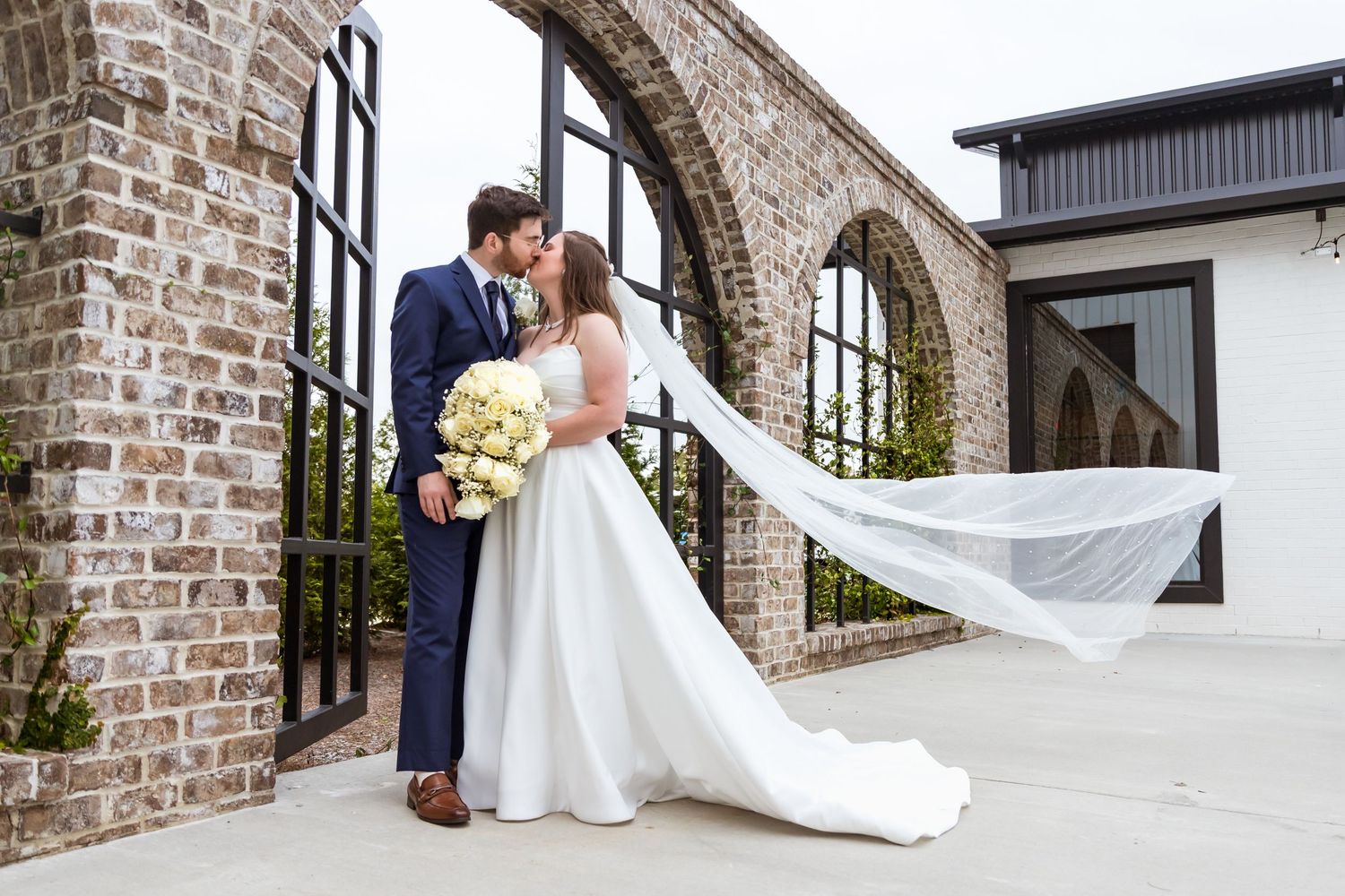 Bride and groom share a kiss outdoors by brick arches, her veil flowing in the wind, holding a white bouquet.