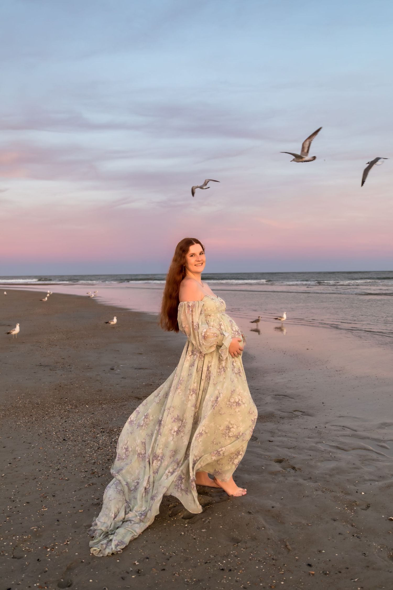 Pregnant woman in floral dress on beach at sunset with seagulls flying overhead.