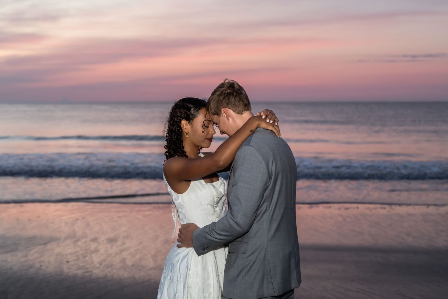 Newlywed couple embracing on beach at sunset, bride in white dress and groom in gray suit.