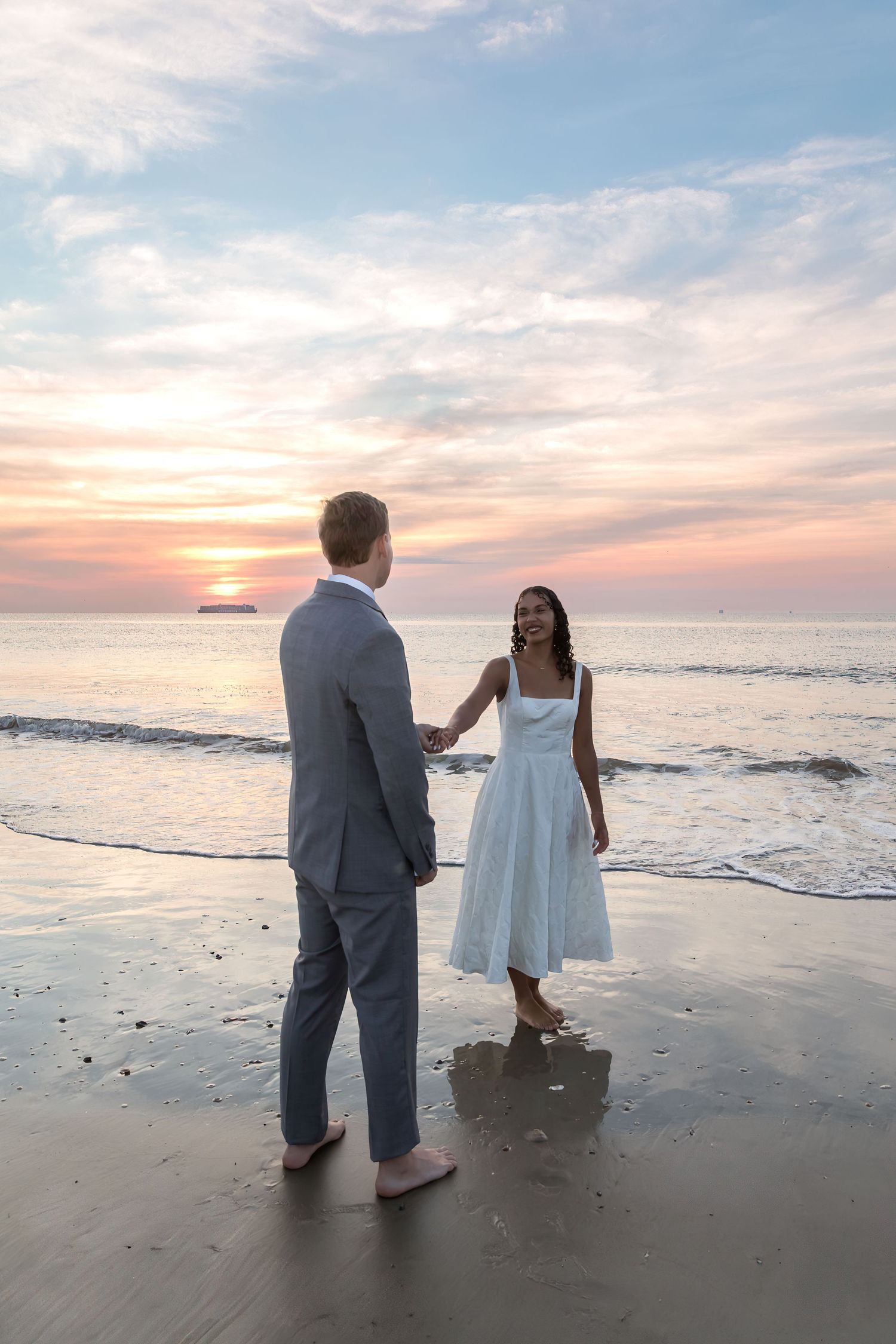 Couple holding hands on beach at sunset, woman in white dress and man in gray suit, waves in background.