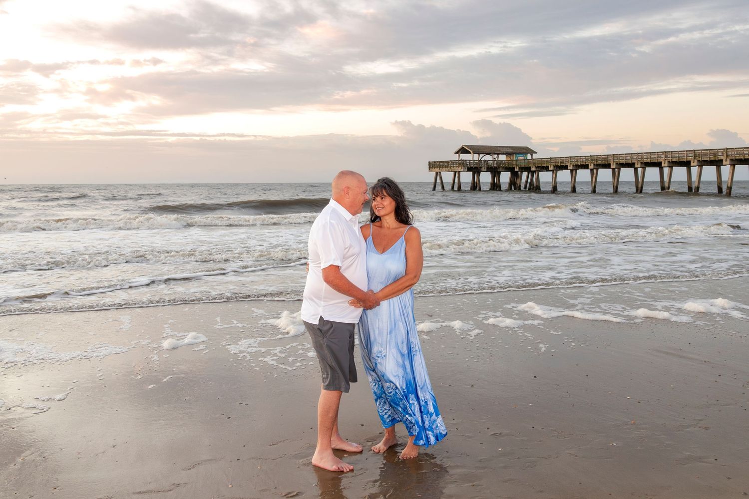 Couple embracing on beach at sunset with pier in background.