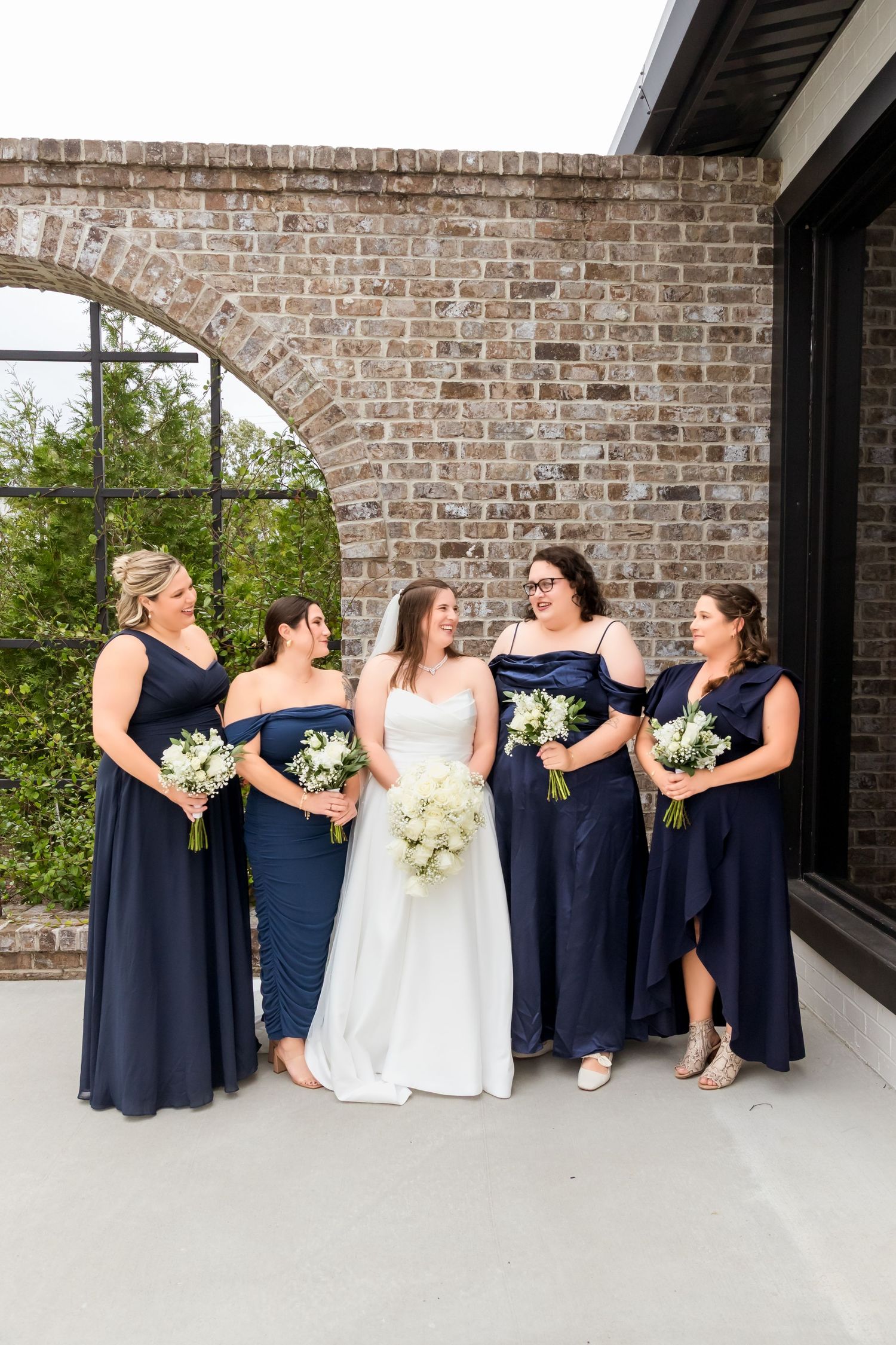 Bridesmaids in navy dresses share candid laughing moments against brick wall backdrop.