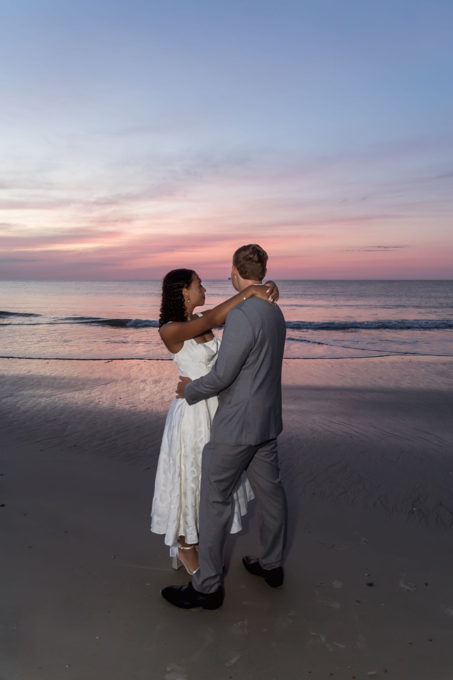Couple in wedding attire dancing romantically on a beach at sunset with pink and purple sky.