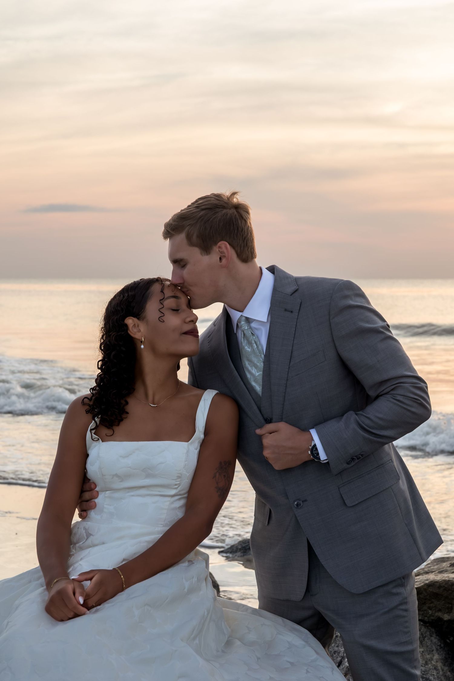 Bride and groom share a romantic kiss on rocky beach at sunset, groom in gray suit, bride in white gown.