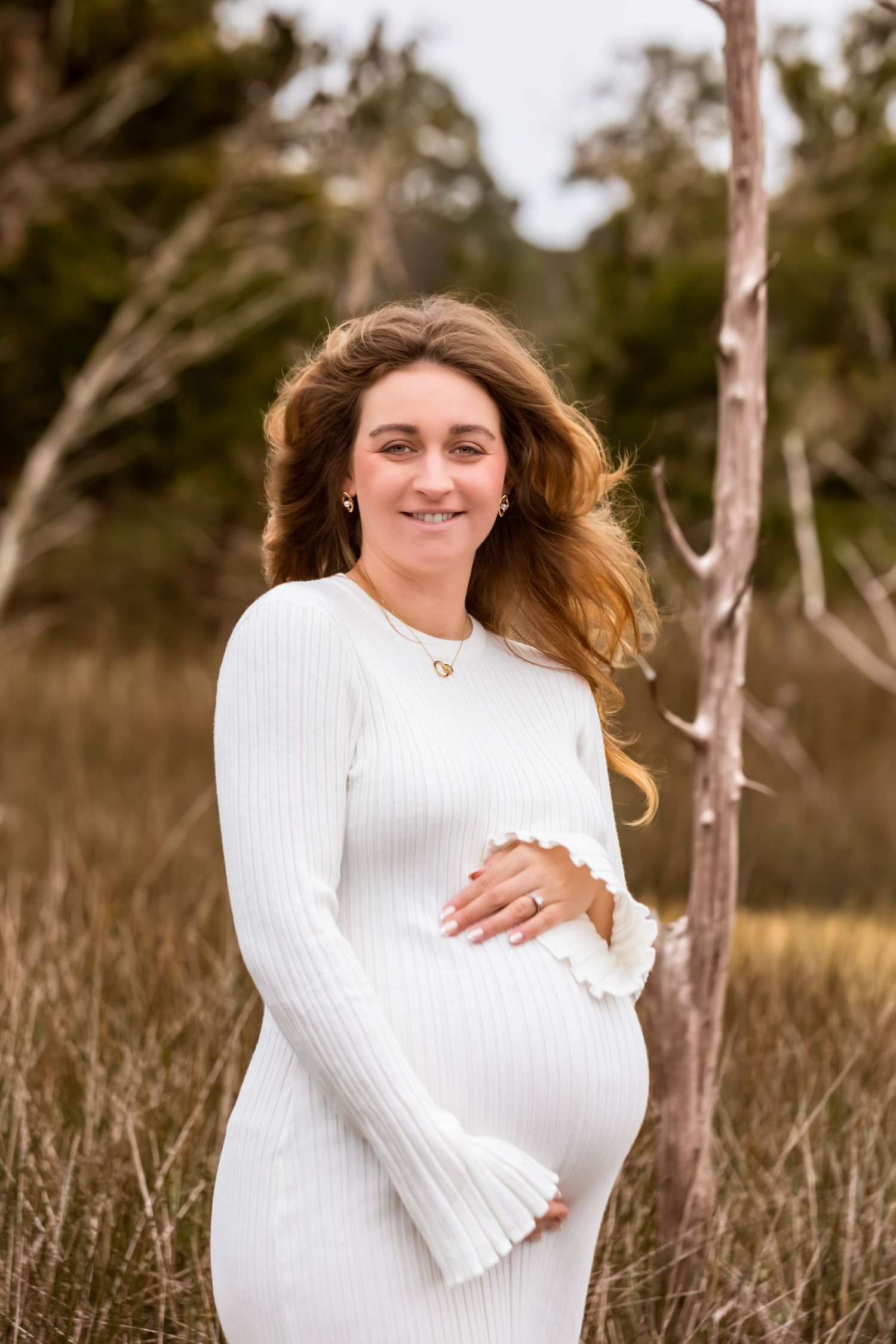 Expectant woman in white ribbed dress poses outdoors in a natural setting with long flowing hair.