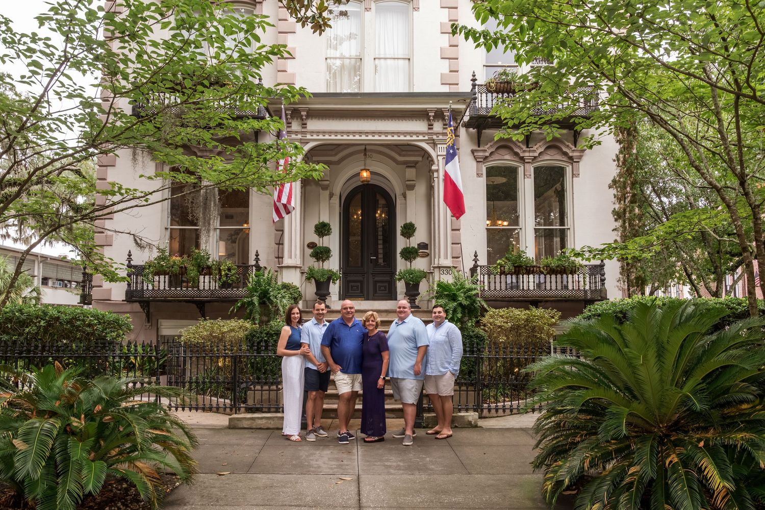Group of people posing in front of an elegant white Southern mansion with flags and lush greenery.