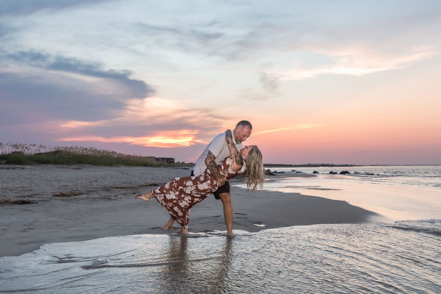 Couple sharing a romantic dip kiss on a beach at sunset, waves lapping at their feet under a colorful sky.