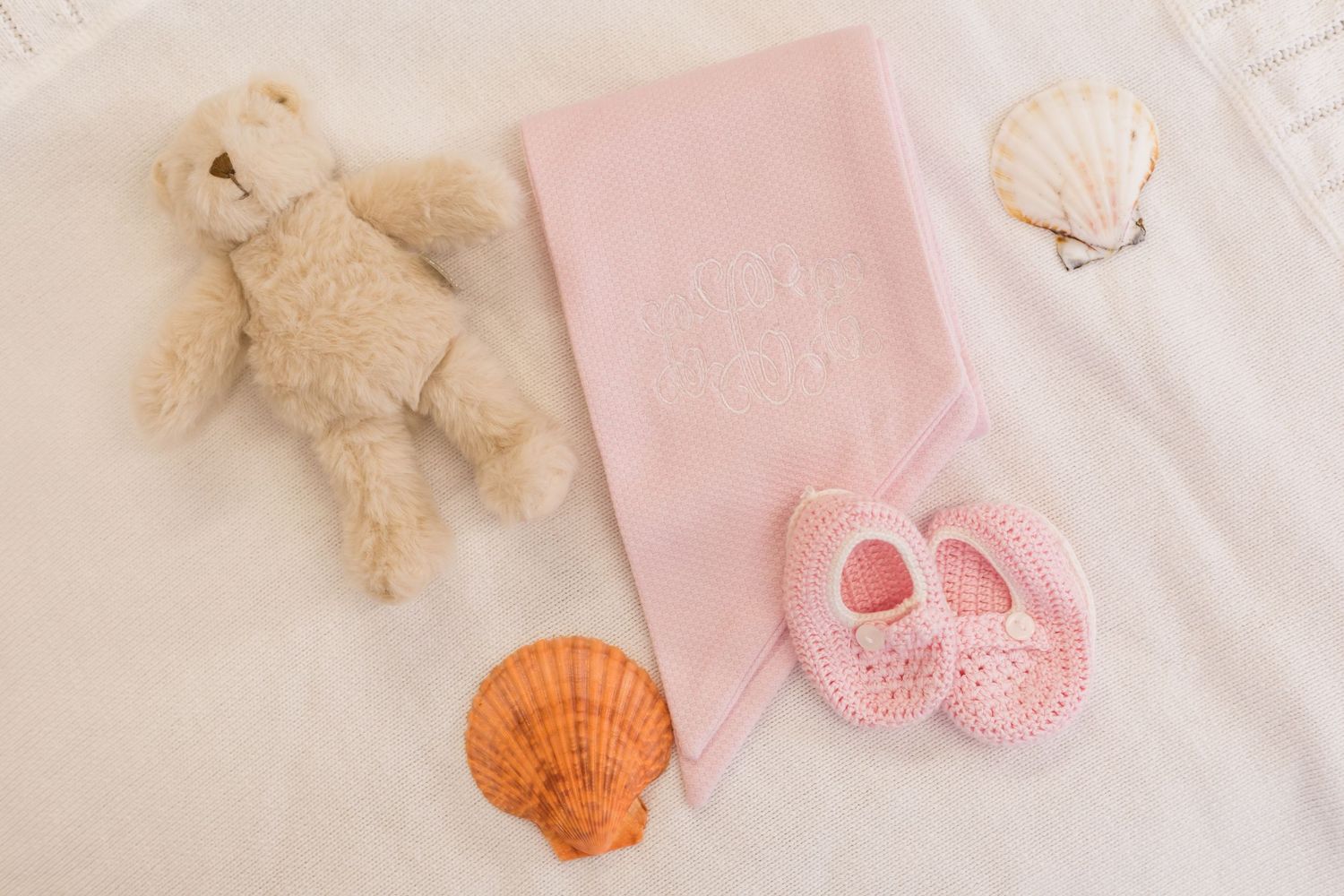 Pink baby blanket with crochet booties, teddy bear, and seashells on white background.