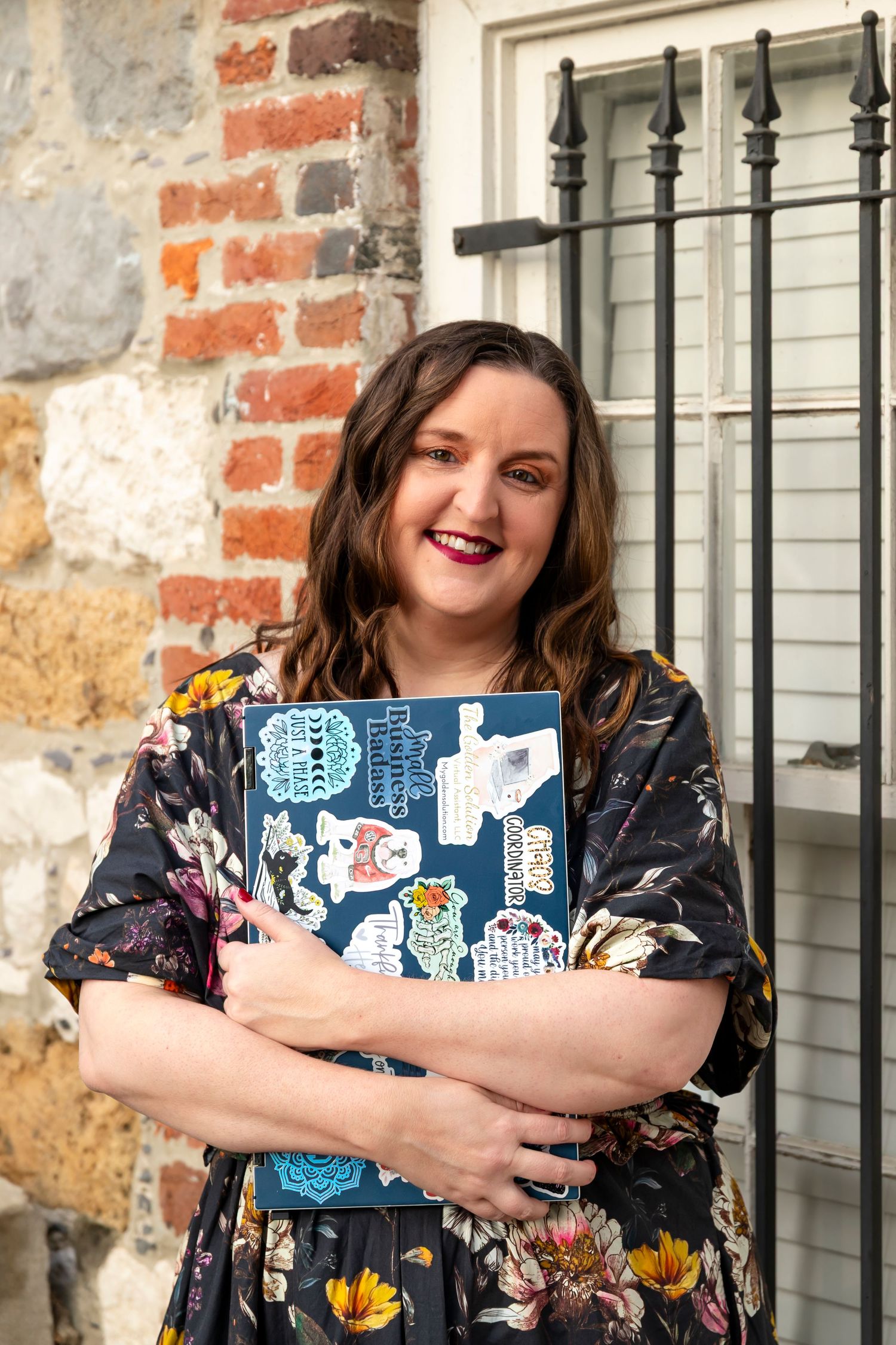 A person in a floral dress holds a decorated laptop against a brick wall exterior.