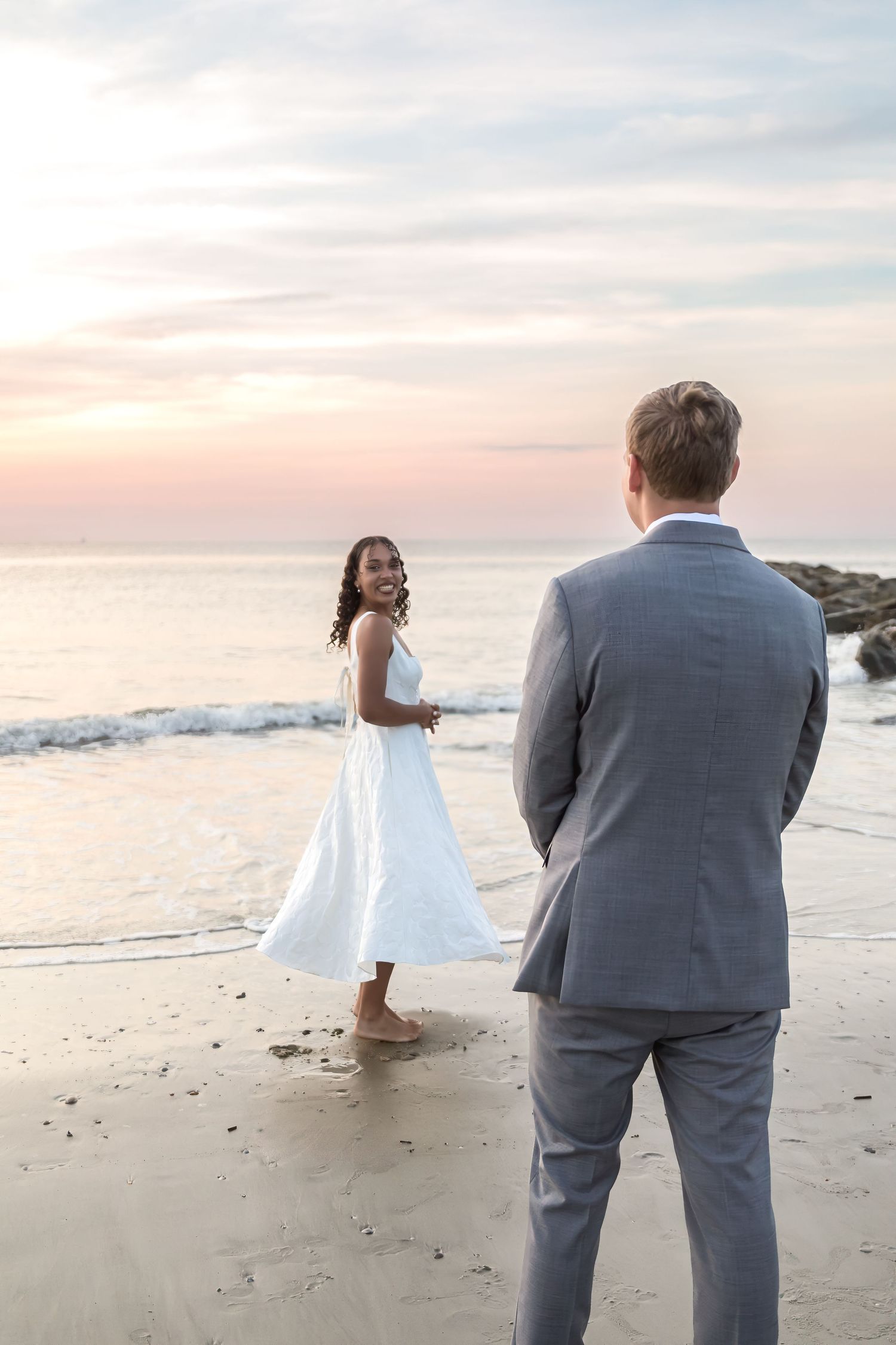 Bride in white dress smiles at groom in gray suit during romantic beach wedding at sunset.