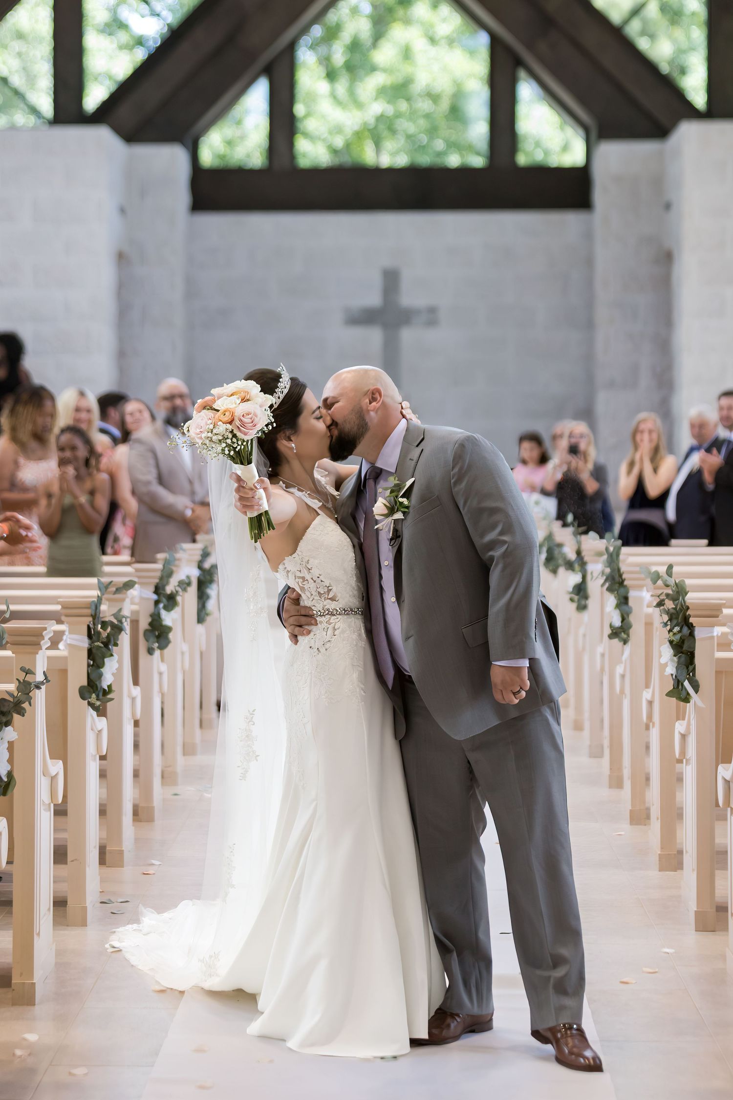 Bride and groom share first kiss at church altar, bride holding bouquet, groom in gray suit, guests applauding in pews.