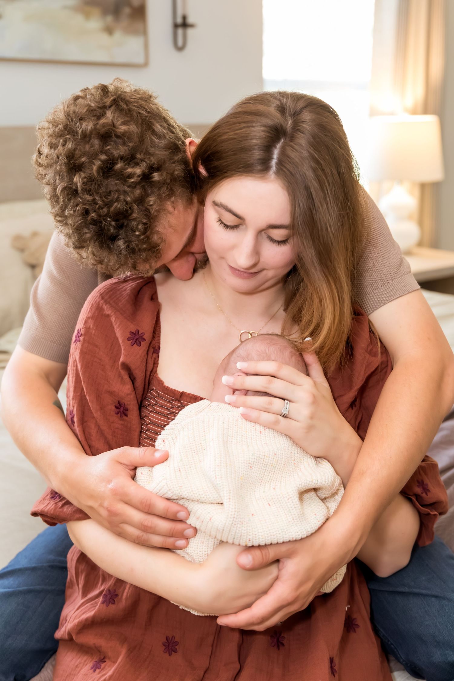 New parents lovingly cradle their newborn together, father kissing mothers head in cozy bedroom setting.