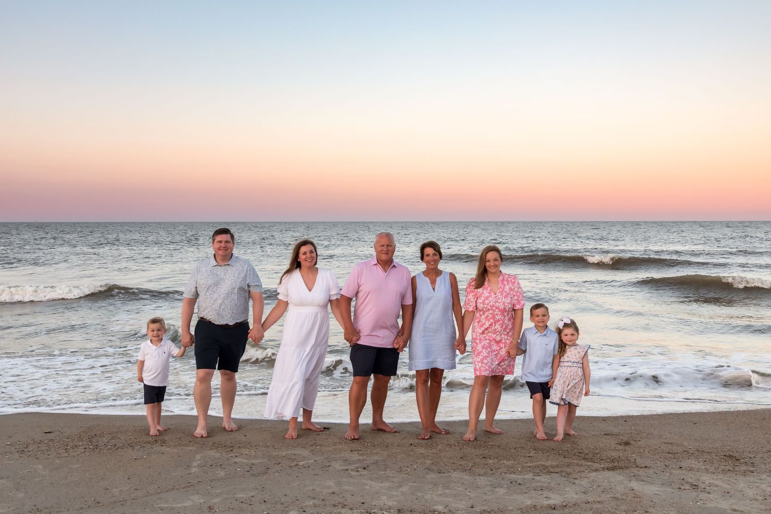 Family of seven poses on a sandy beach at sunset, with ocean waves and a pink and purple sky behind them.