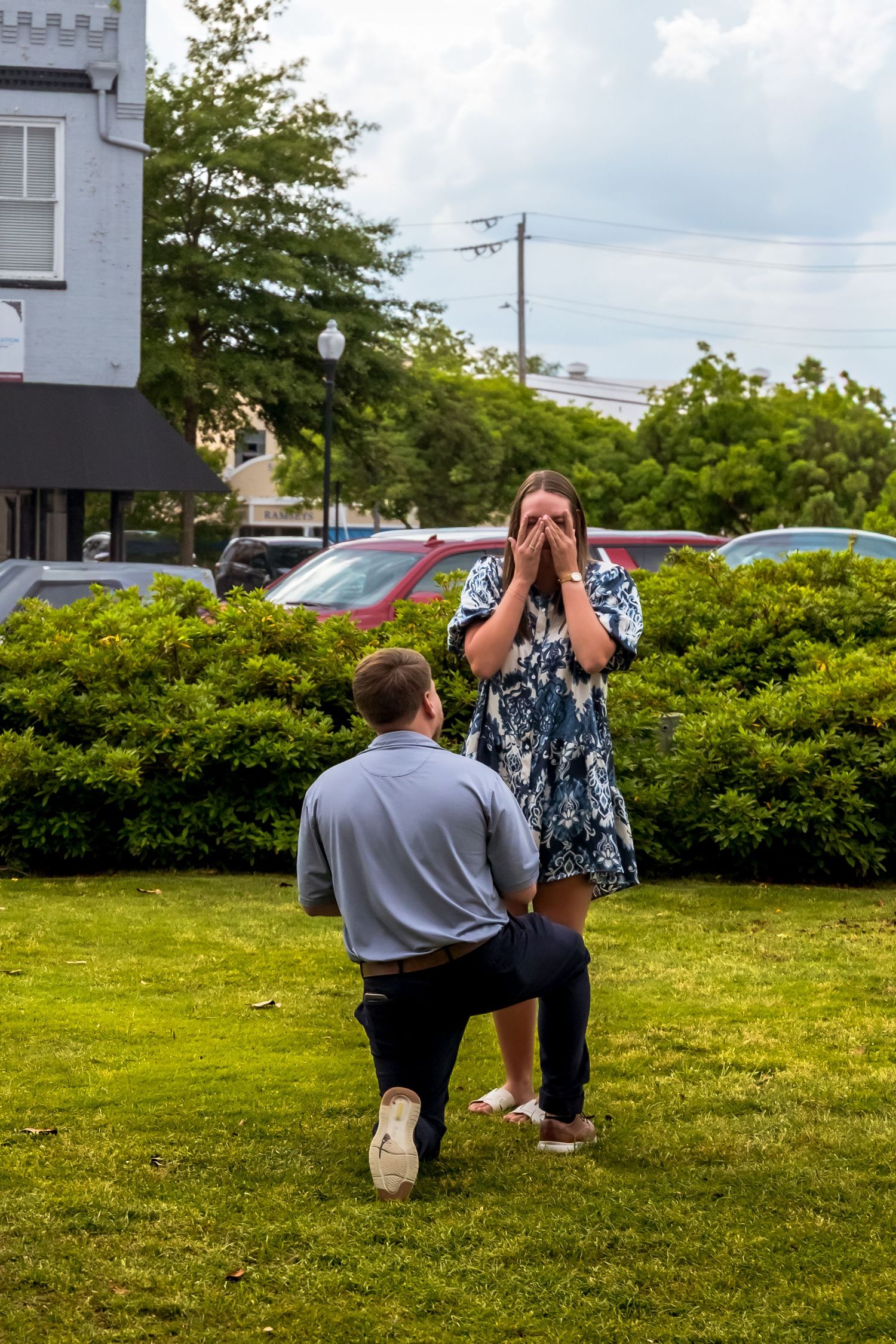 Couple kneeling in proposal position on grassy lawn near building.