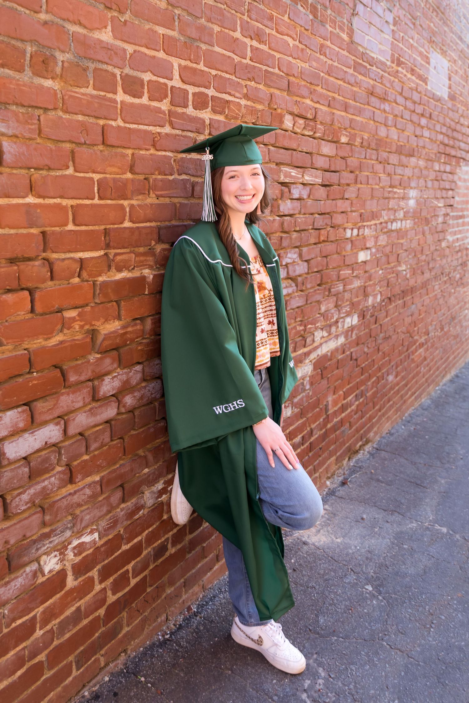 Smiling graduate in green cap and gown leaning against red brick wall wearing jeans and white sneakers.