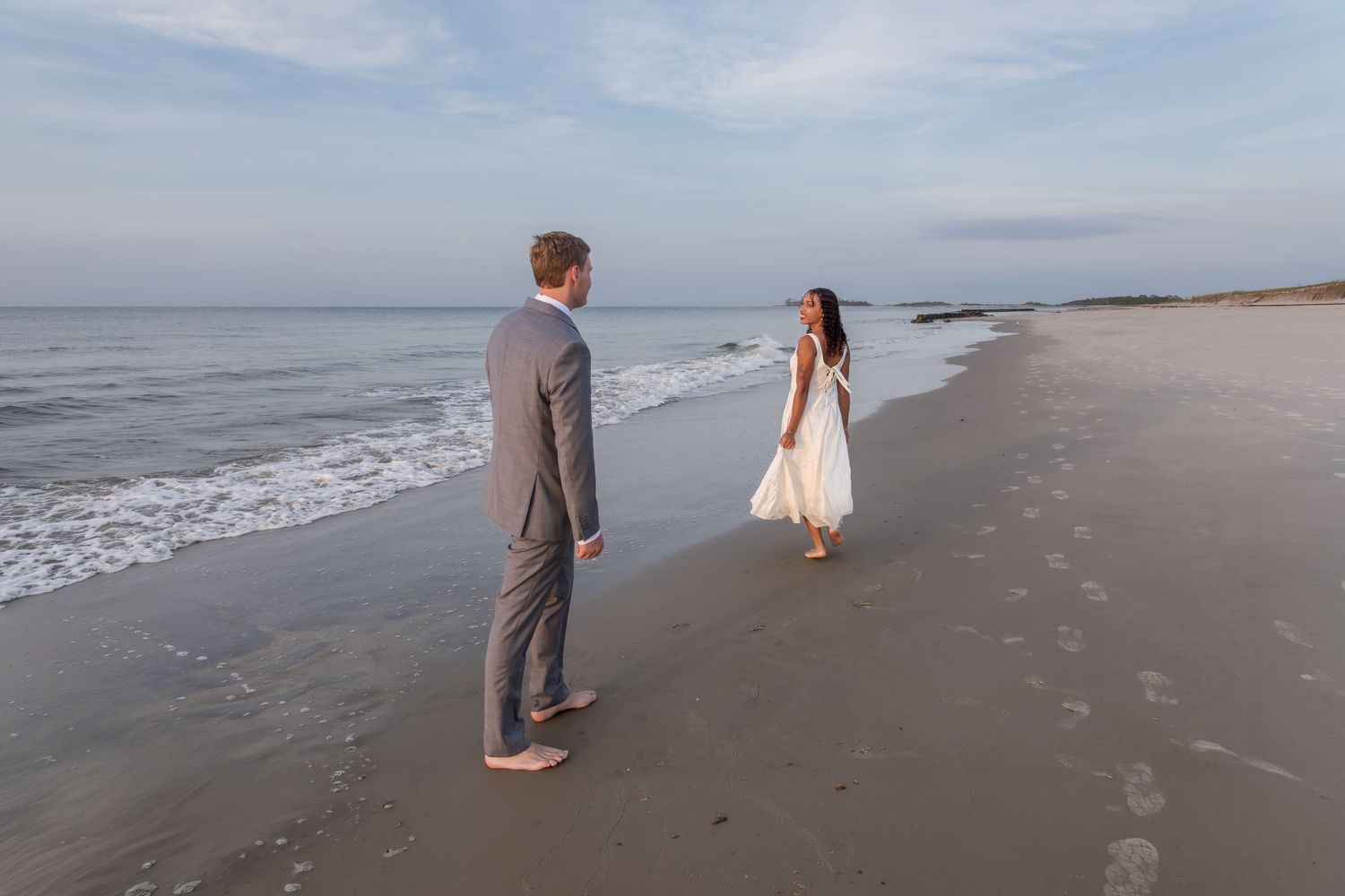 Barefoot couple in wedding attire face each other on sandy beach at dusk.