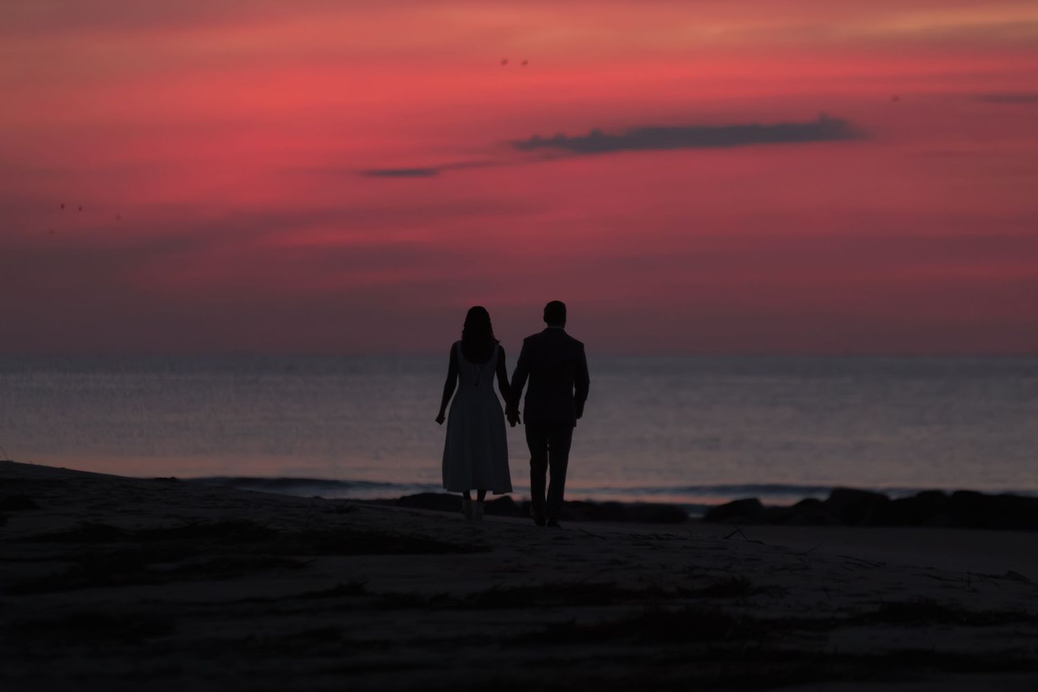Silhouette of a couple holding hands on a beach at sunset with a vibrant red and pink sky over the ocean.