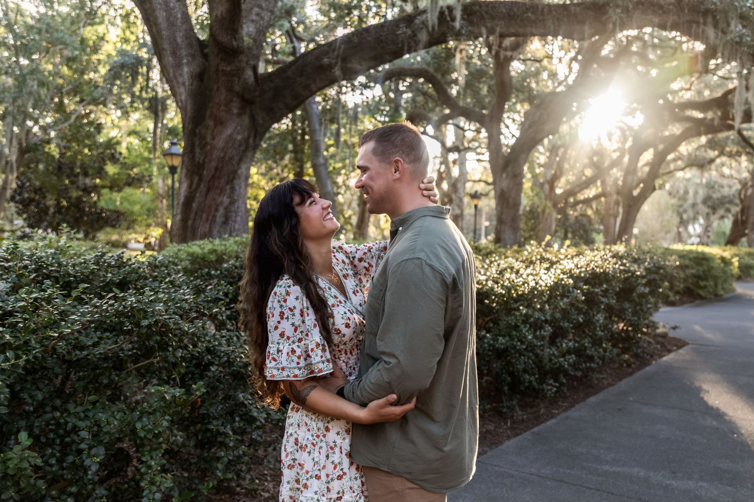 Couple embracing and smiling at each other on a sunlit path surrounded by lush green trees and bushes.