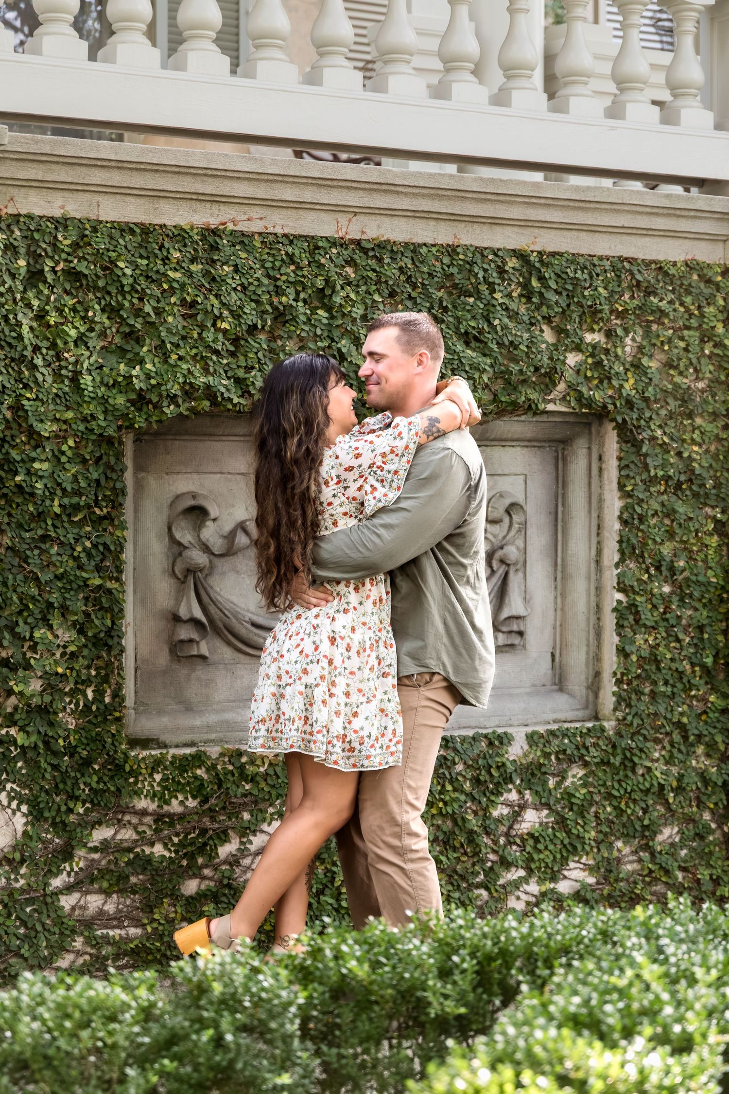 Couple embracing and smiling at each other in a lush ivy-covered garden wall with ornate stone decor.
