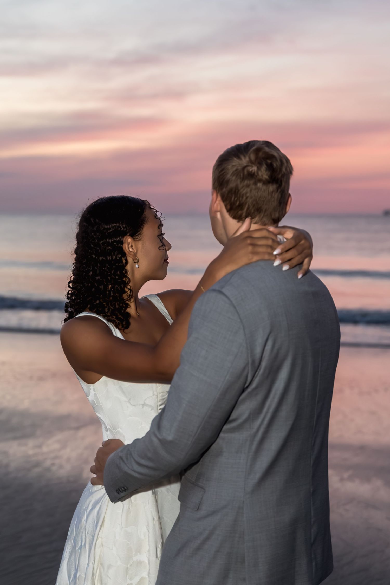 Couple in wedding attire embracing on beach at sunset, bride in white dress and groom in gray suit.
