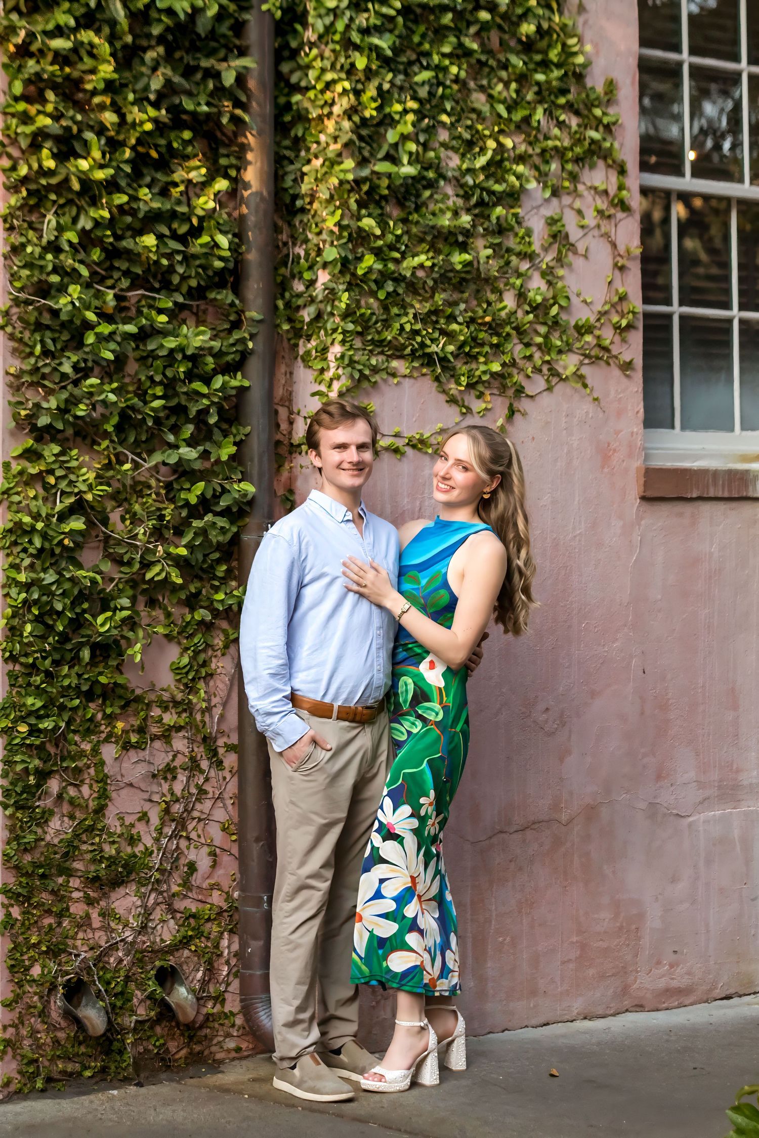 A couple poses romantically against an ivy-covered pink wall, woman in floral green dress, man in light blue shirt.