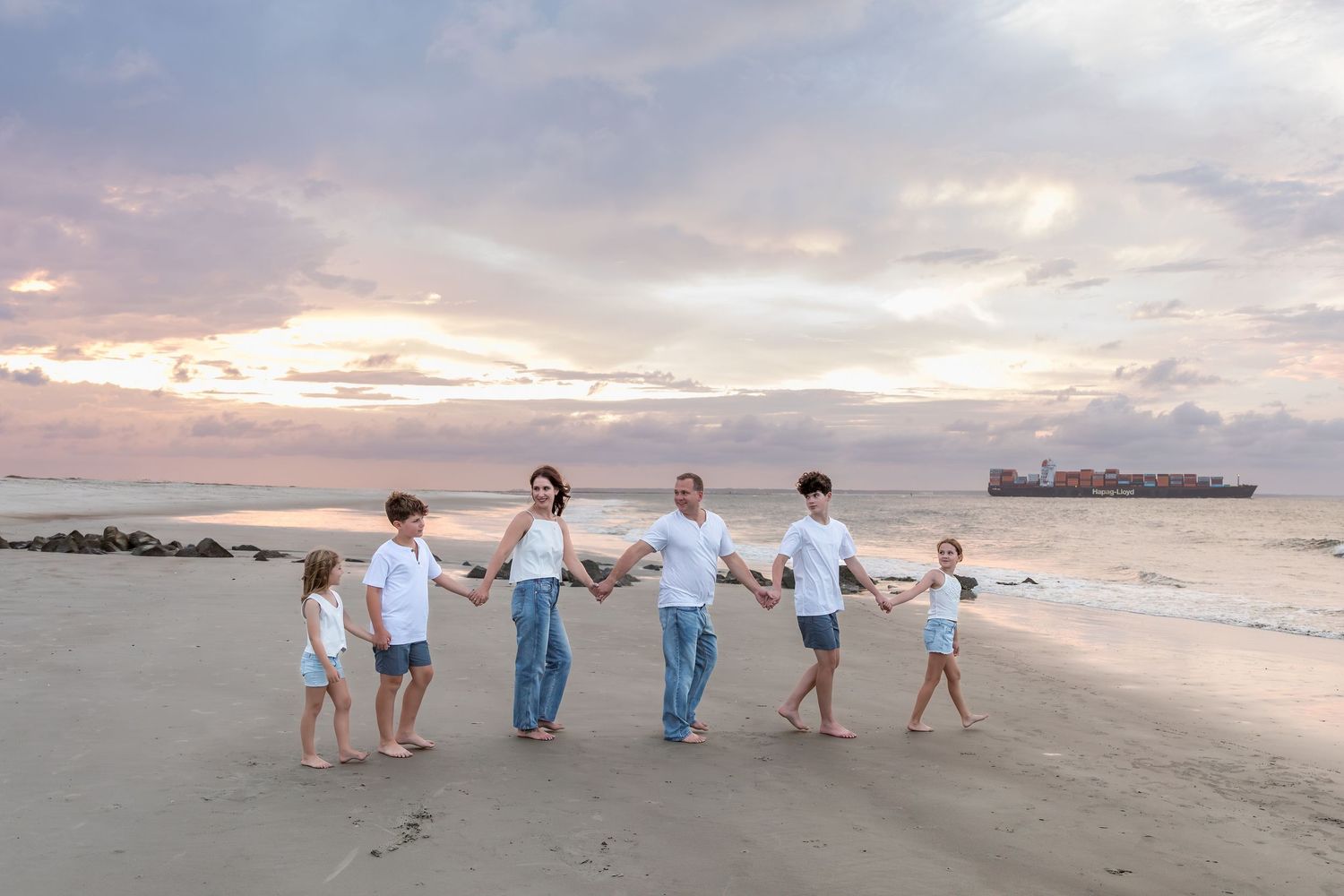 Family of six holding hands on a beach at sunset, wearing white tops and jeans, with ocean waves and colorful sky.