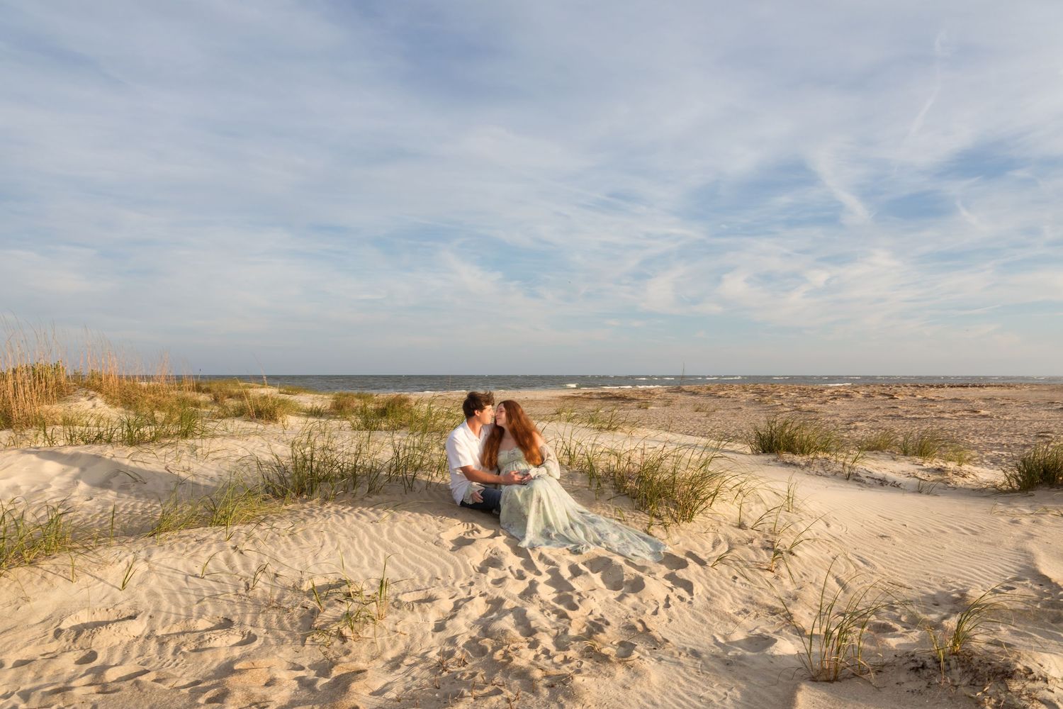 A couple shares a romantic kiss on sandy beach dunes with ocean views and wispy clouds in the background.