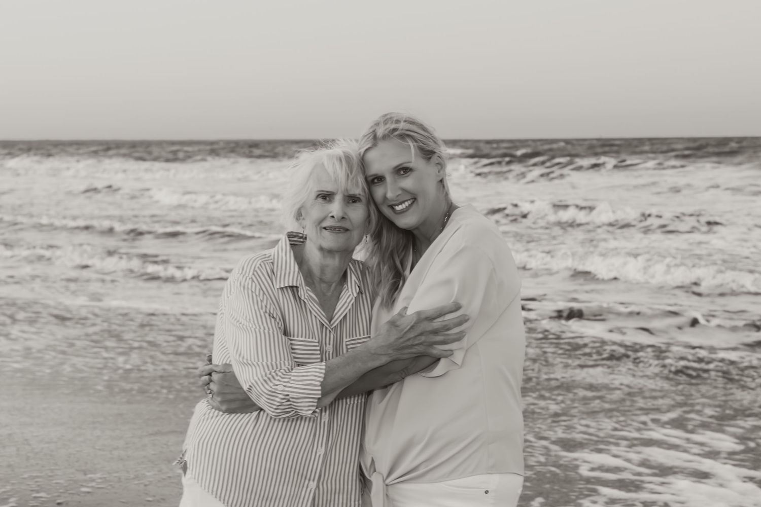 Two women embrace and smile on a beach with ocean waves, captured in a classic black and white portrait photo.