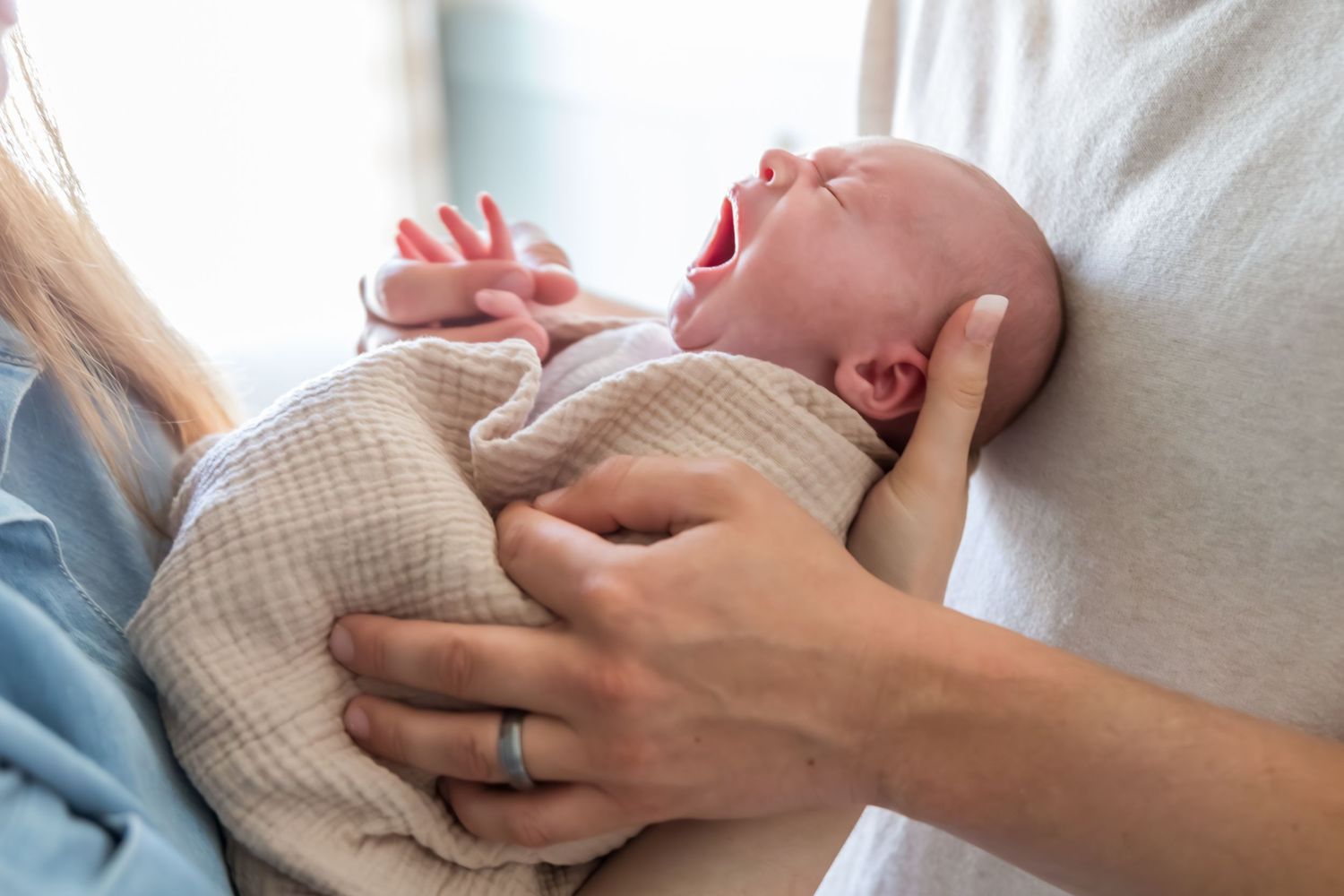 A newborn baby yawning while being cradled in a parent's arms, wrapped in a soft pink blanket.