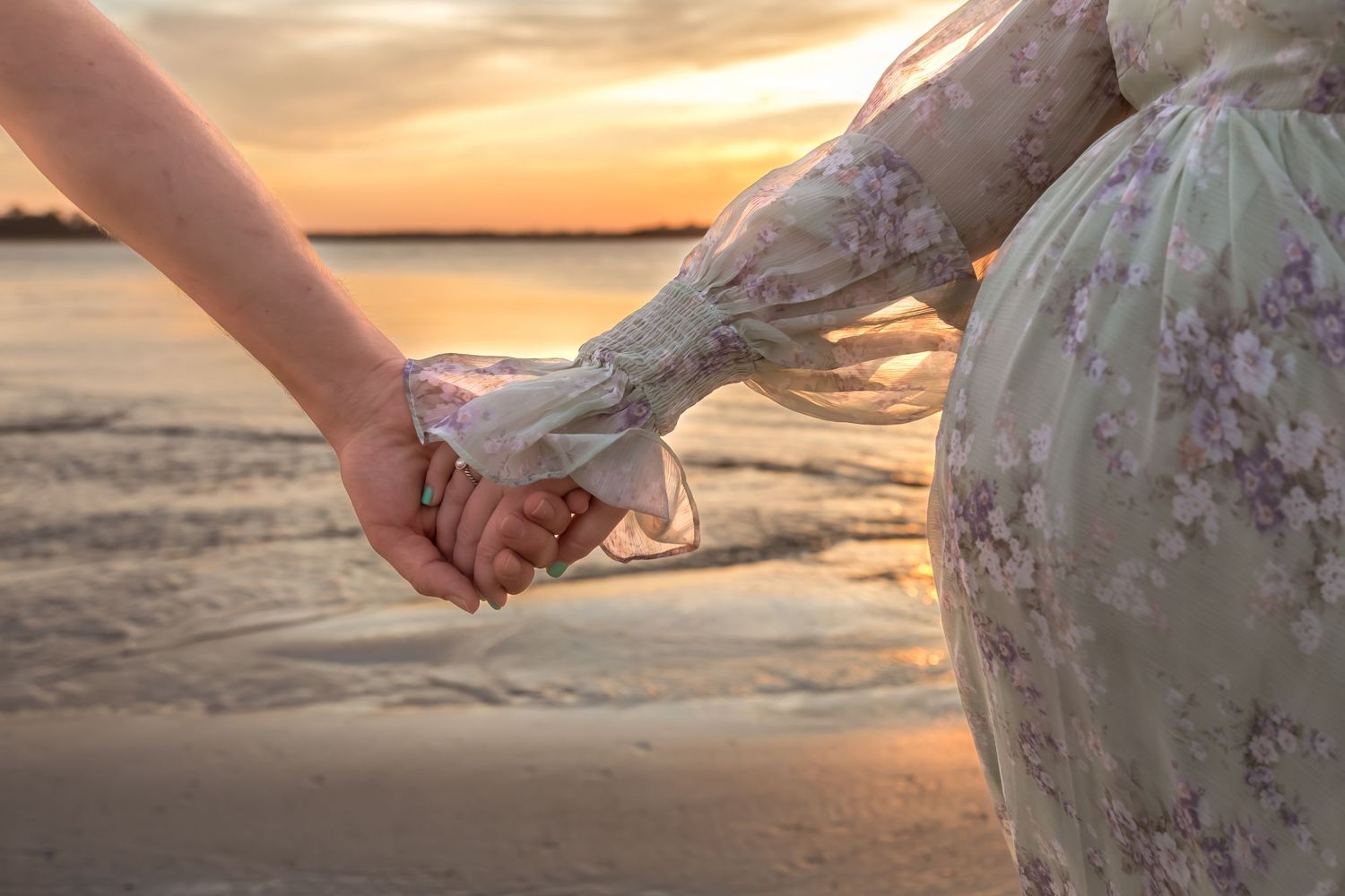 Two people holding hands at sunset on a beach, one wearing a floral green dress, romantic golden light.