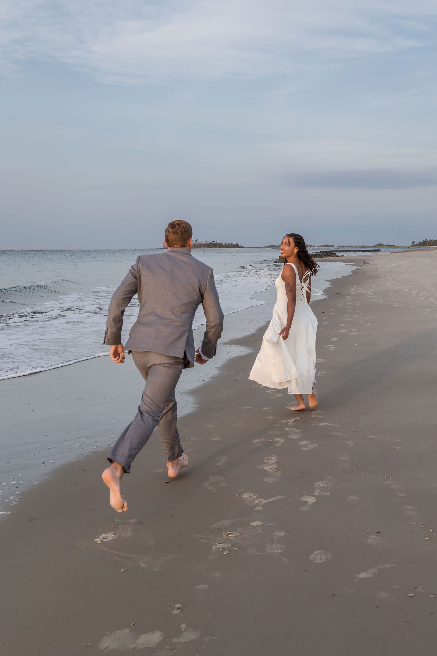 Barefoot bride in white dress and groom in gray suit running on sandy beach at dusk near ocean waves.